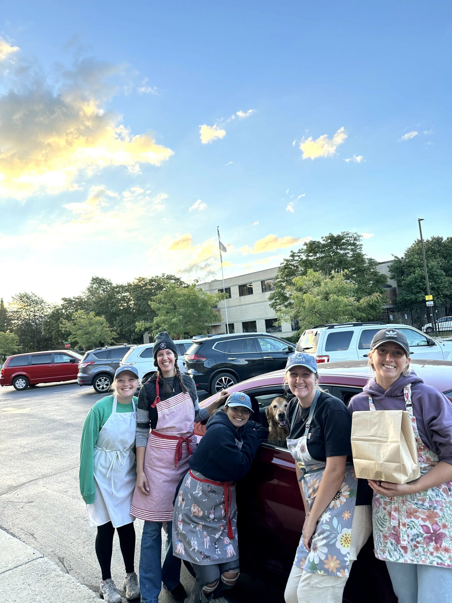 Group of five people standing outdoors in a parking lot with cars and trees, smiling at the camera.