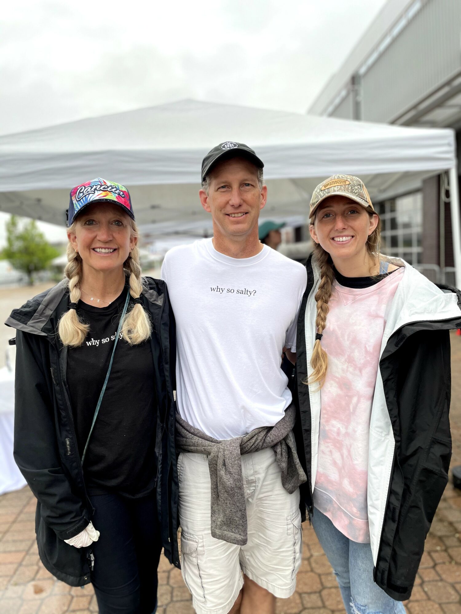 Three people standing outdoors in front of a white tent, smiling, with cloudy sky overhead.
