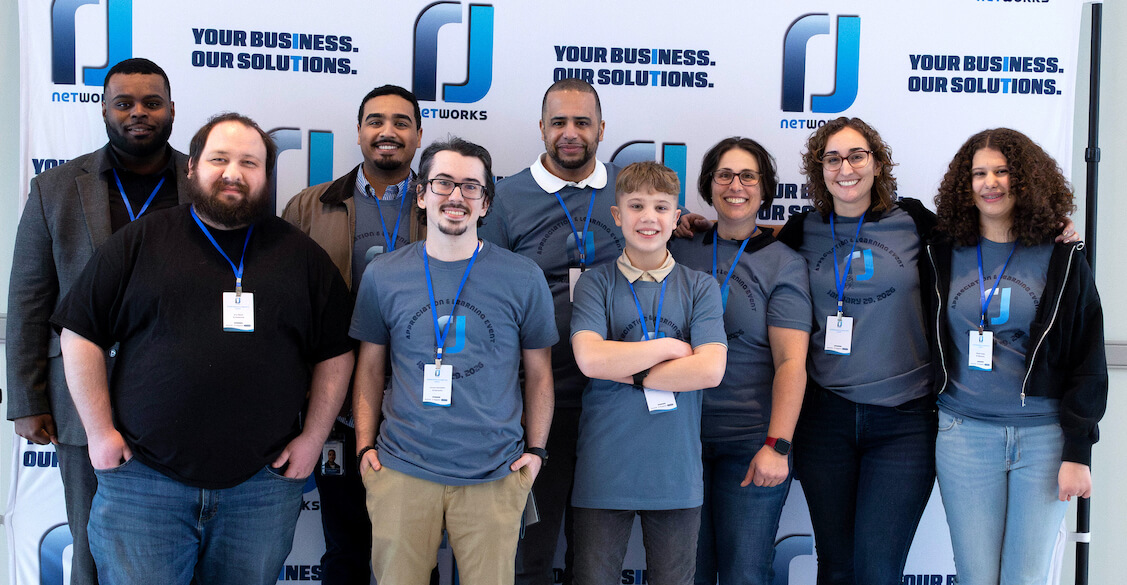 Group of eight people standing in front of a backdrop with logos and text, smiling and wearing name tags.