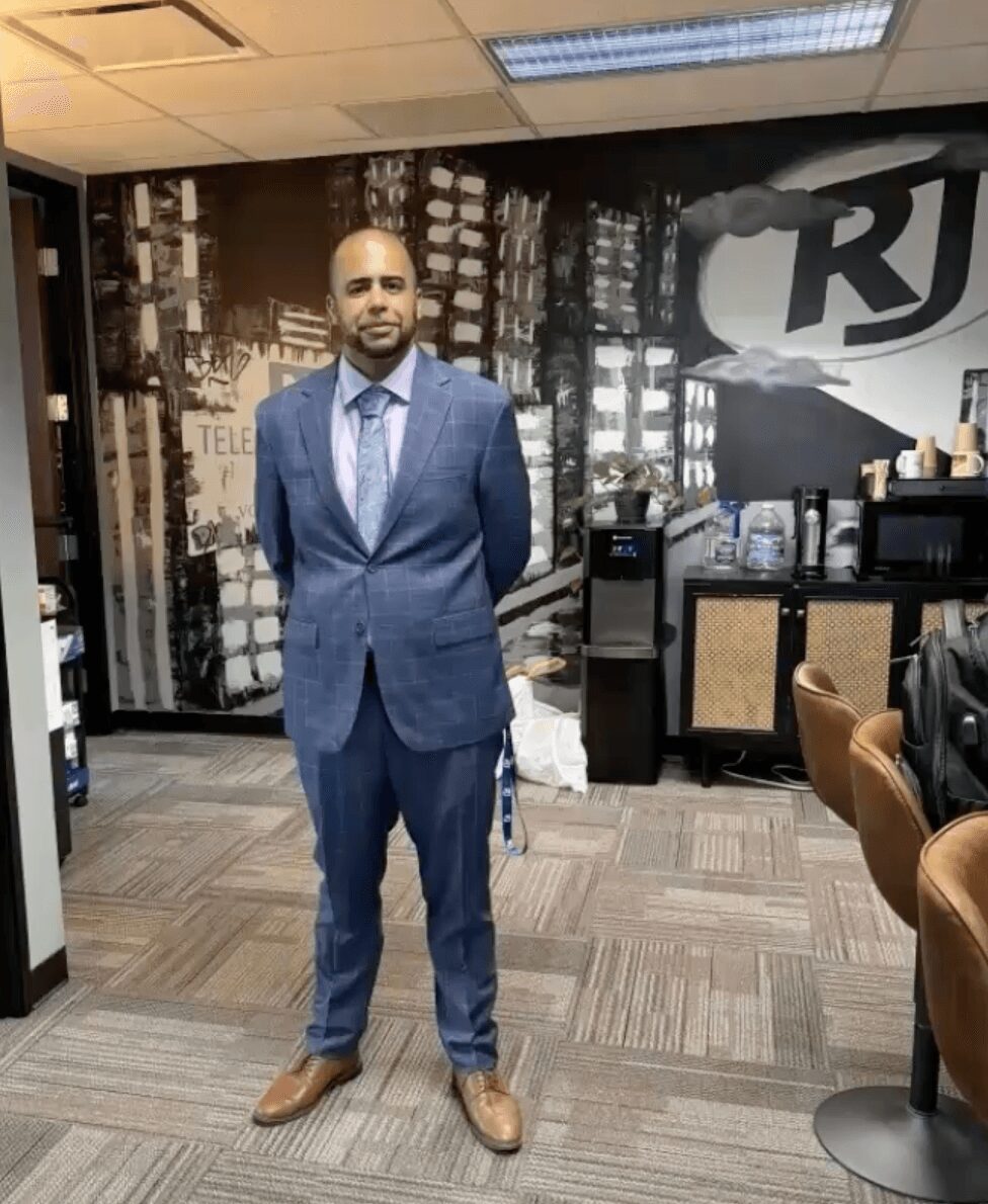 Man in a blue suit standing in an office with a cityscape mural, water cooler, and chairs nearby.