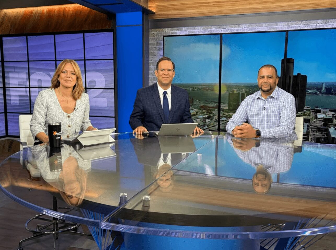 Three news anchors sit at a glass desk in a studio with large screens behind them showing cityscapes.