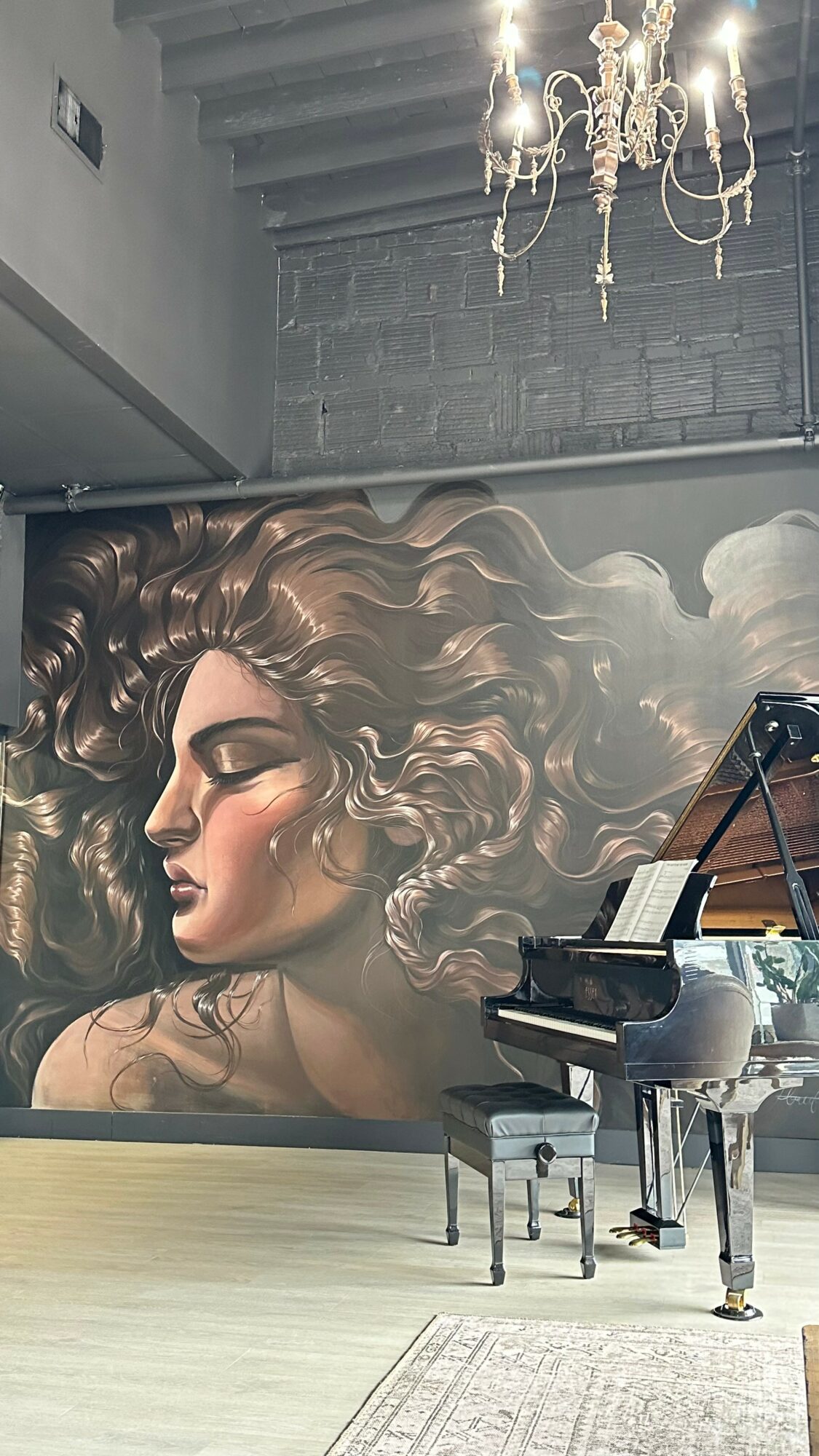 Large mural of a woman with flowing hair on a wall, with a grand piano and chair in front.
