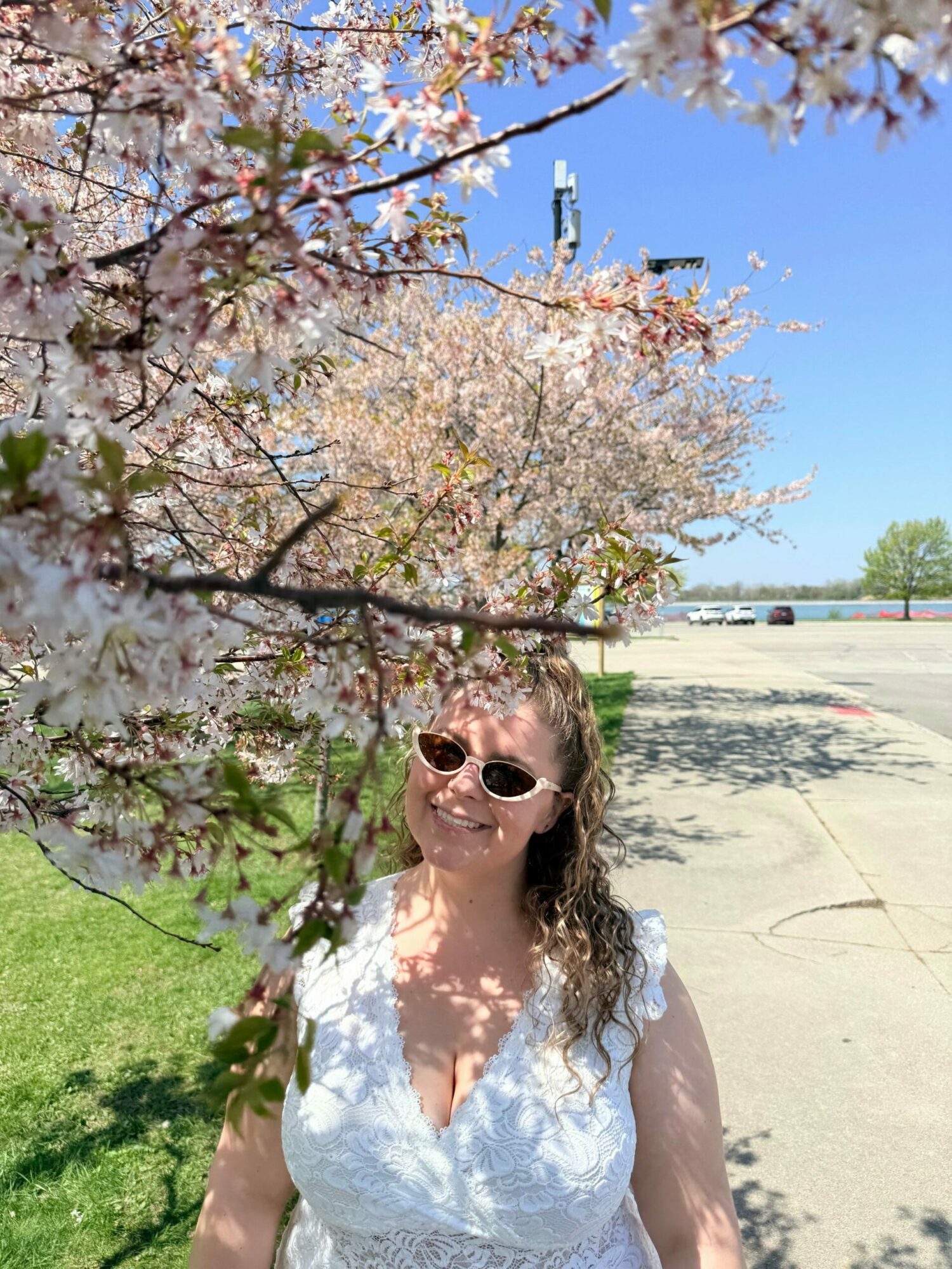 Woman with sunglasses standing near blooming cherry blossom tree in park on sunny day.