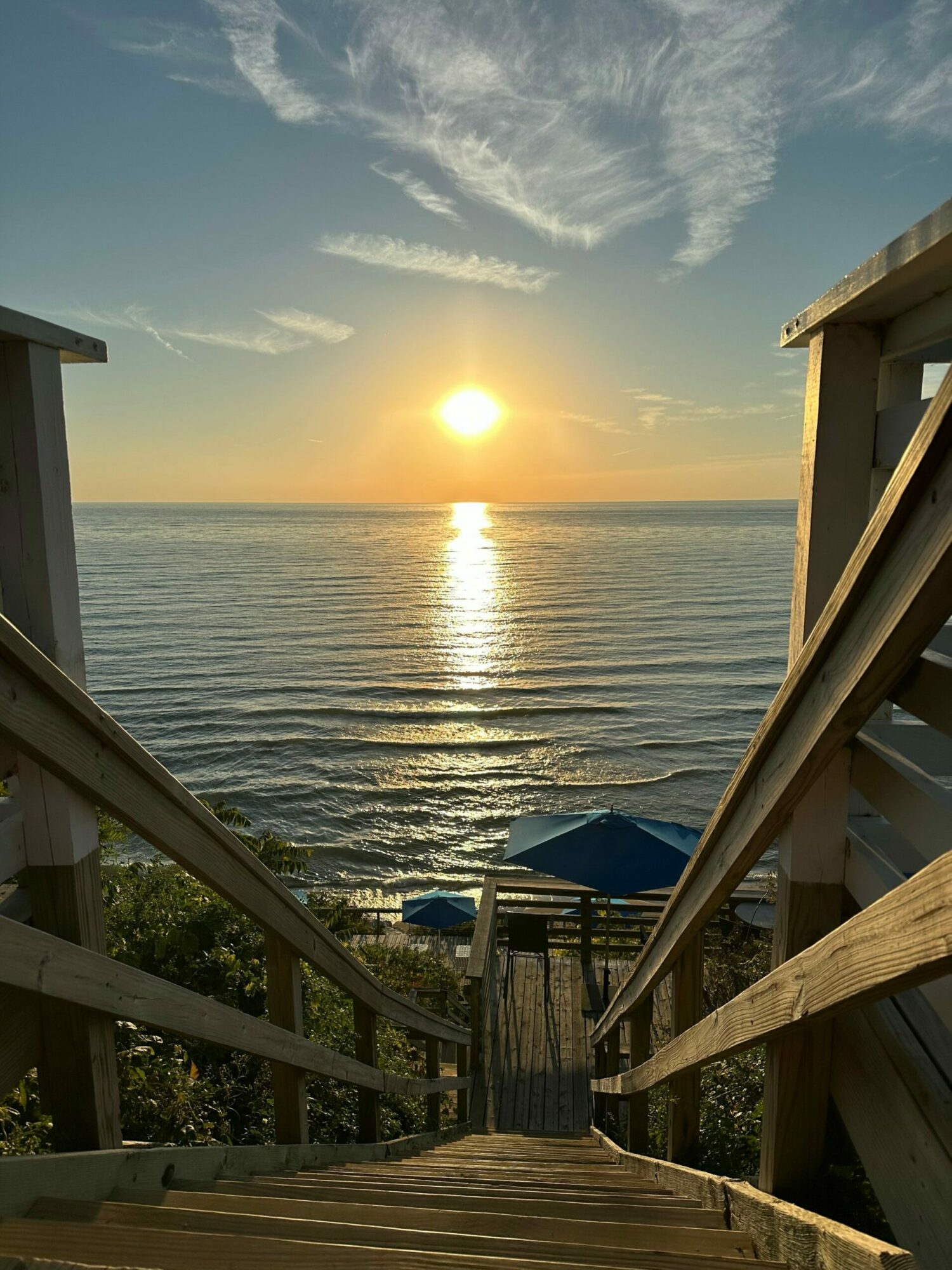 Sunset over calm water viewed from a wooden deck with railings, clouds in the sky, and a distant horizon.