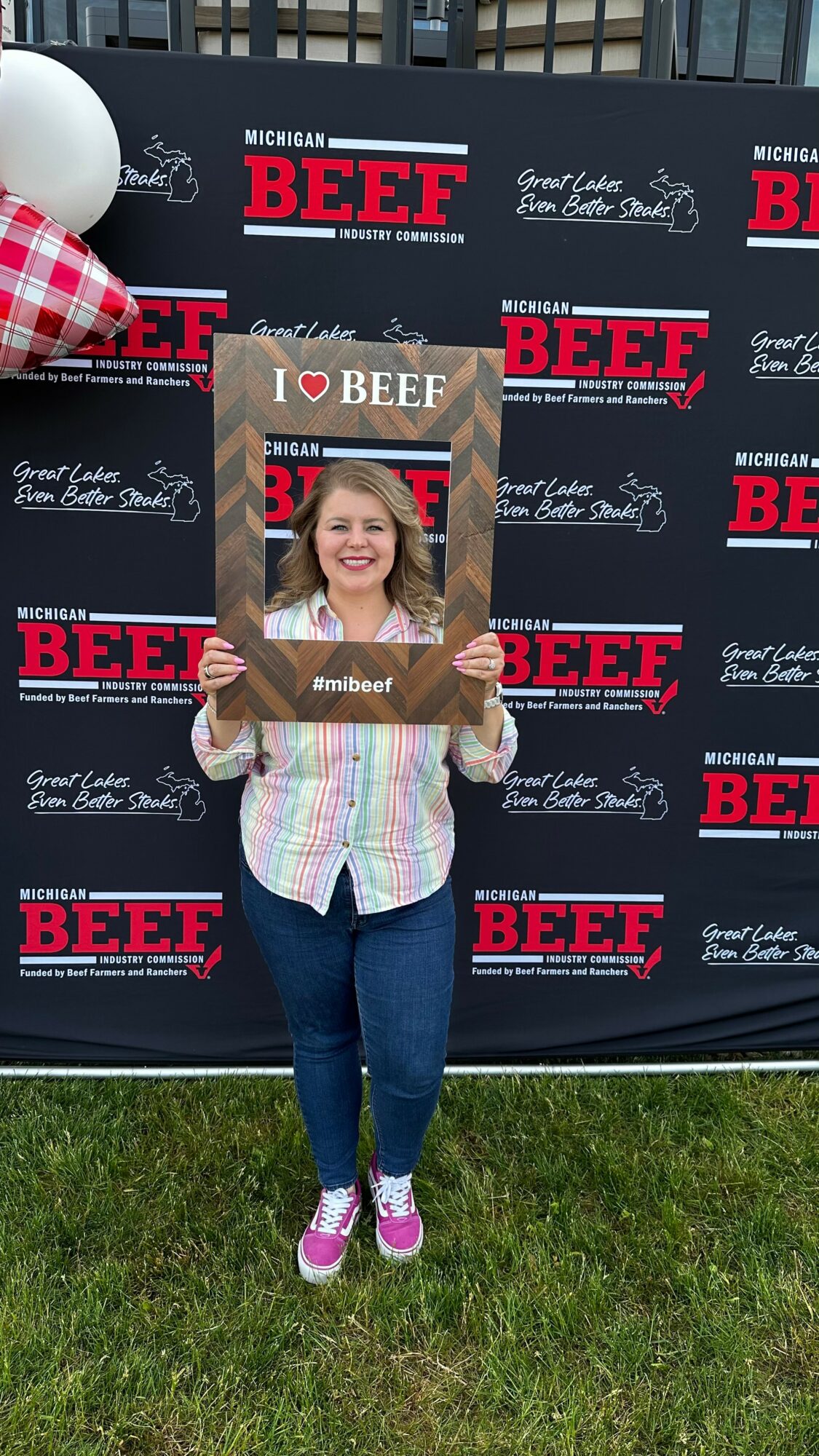 Woman holding a sign in front of a BEEF event backdrop, smiling, wearing a striped shirt and jeans, standing on grass.