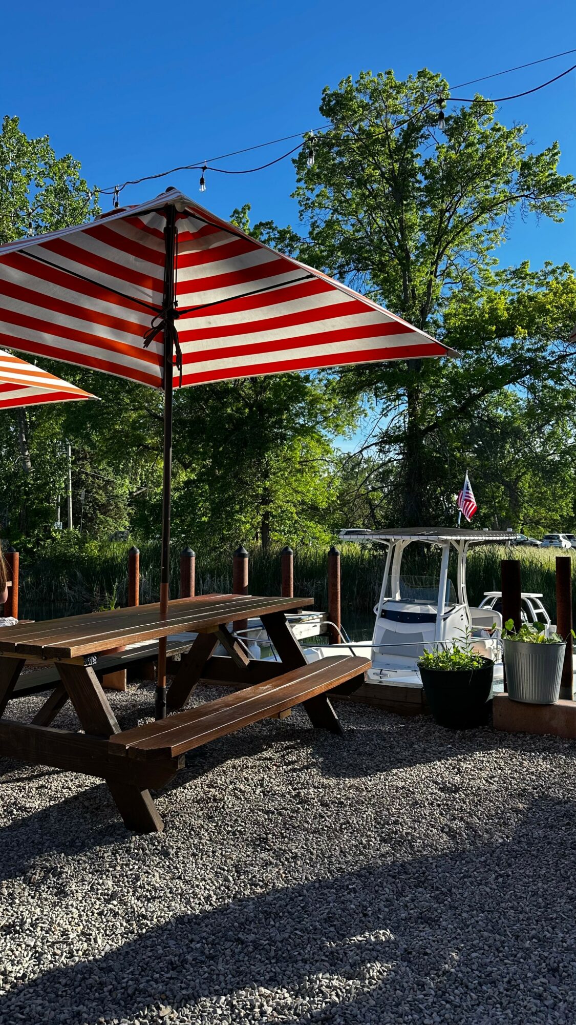 Outdoor seating area with a striped red and white umbrella, wooden picnic table, and potted plants, trees in background, sunny day.