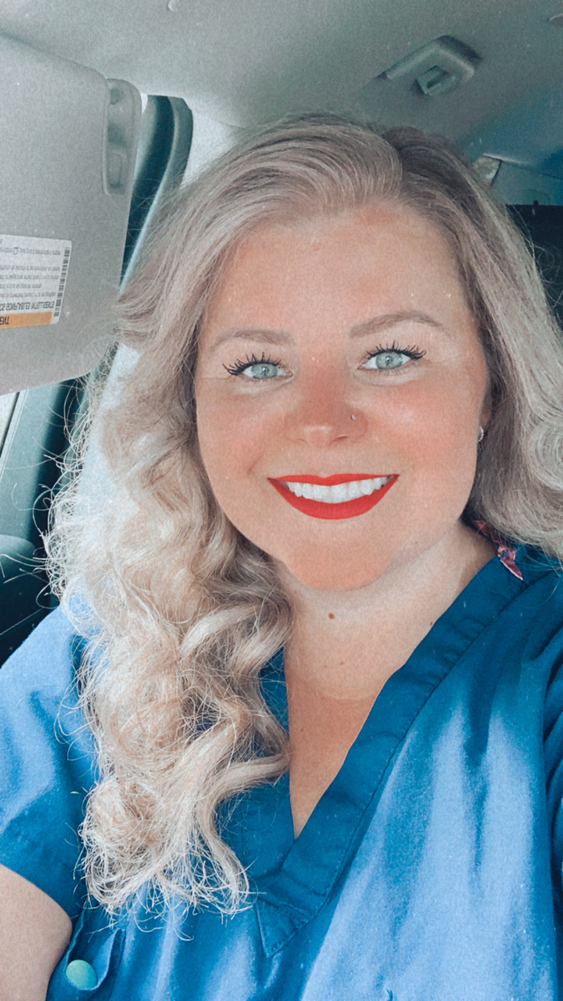 Woman with long, wavy blonde hair smiling, wearing blue scrubs, inside a vehicle.