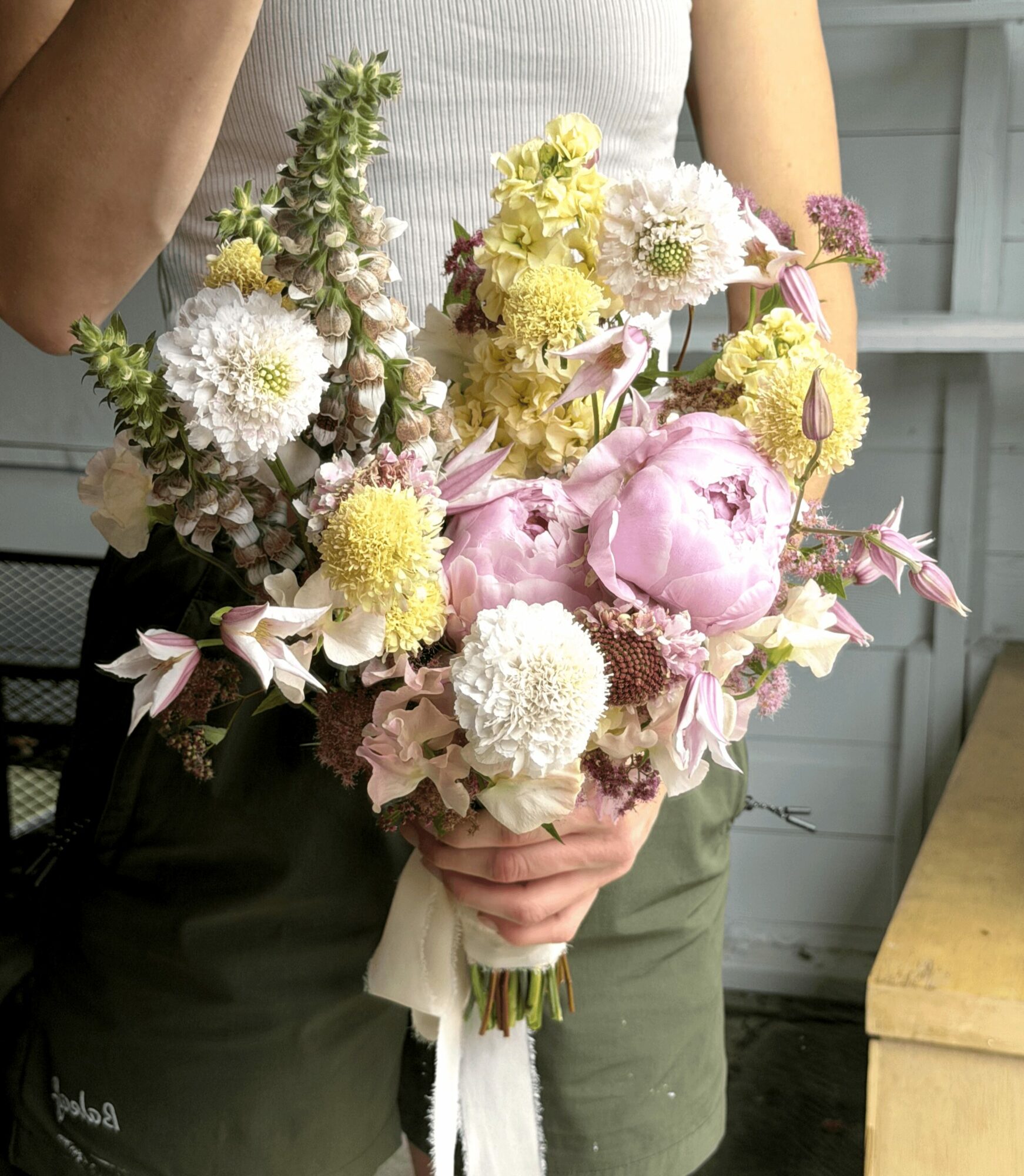 Person holding a bouquet of pink, white, and yellow flowers with a white ribbon, in front of a wooden background.