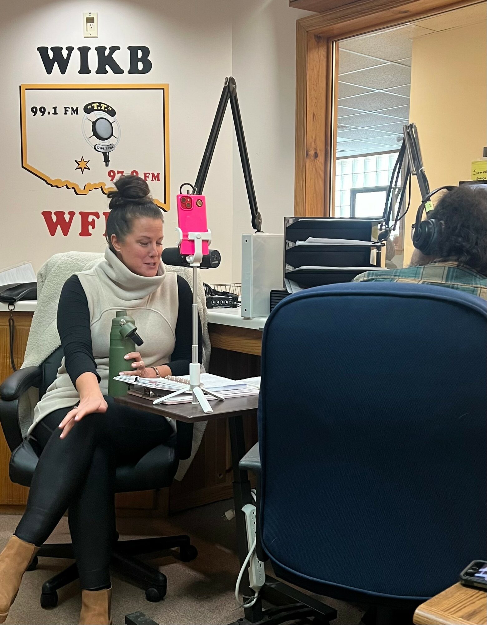 Two women sit in a radio station studio with microphones, headphones, and recording equipment, engaged in conversation.