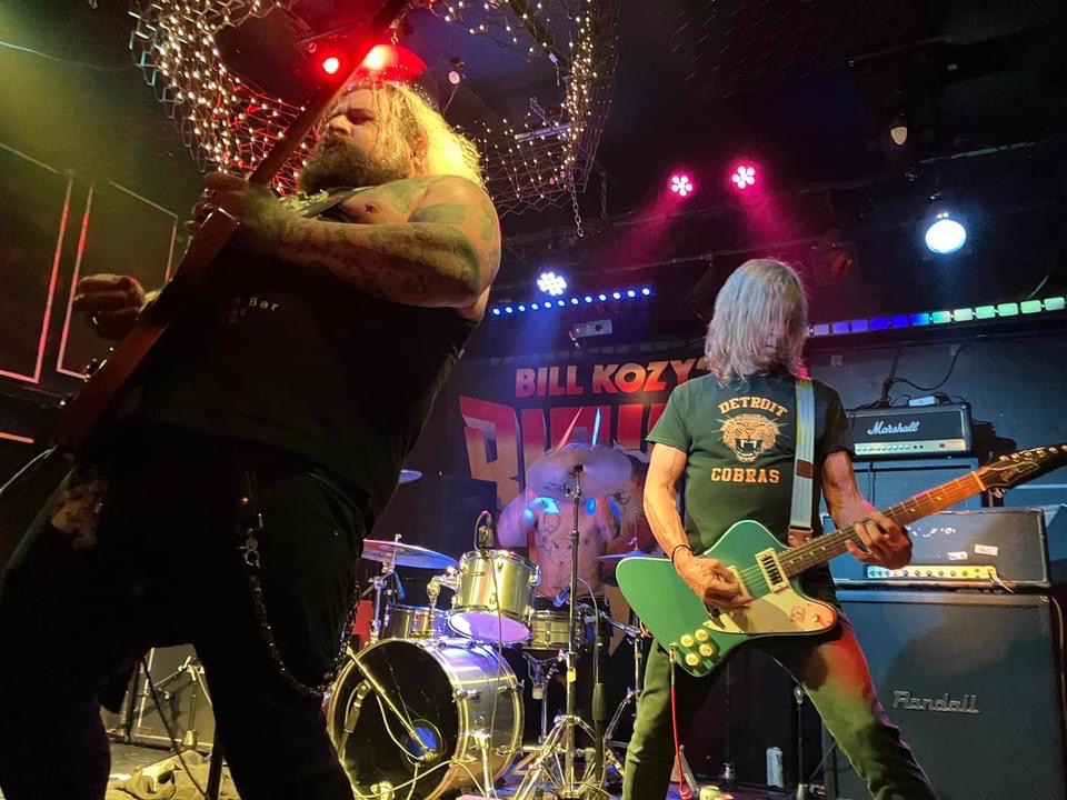 Two musicians perform on stage with drums and amplifiers, colorful lights, and a backdrop with text. One plays guitar, the other stands with a guitar.
