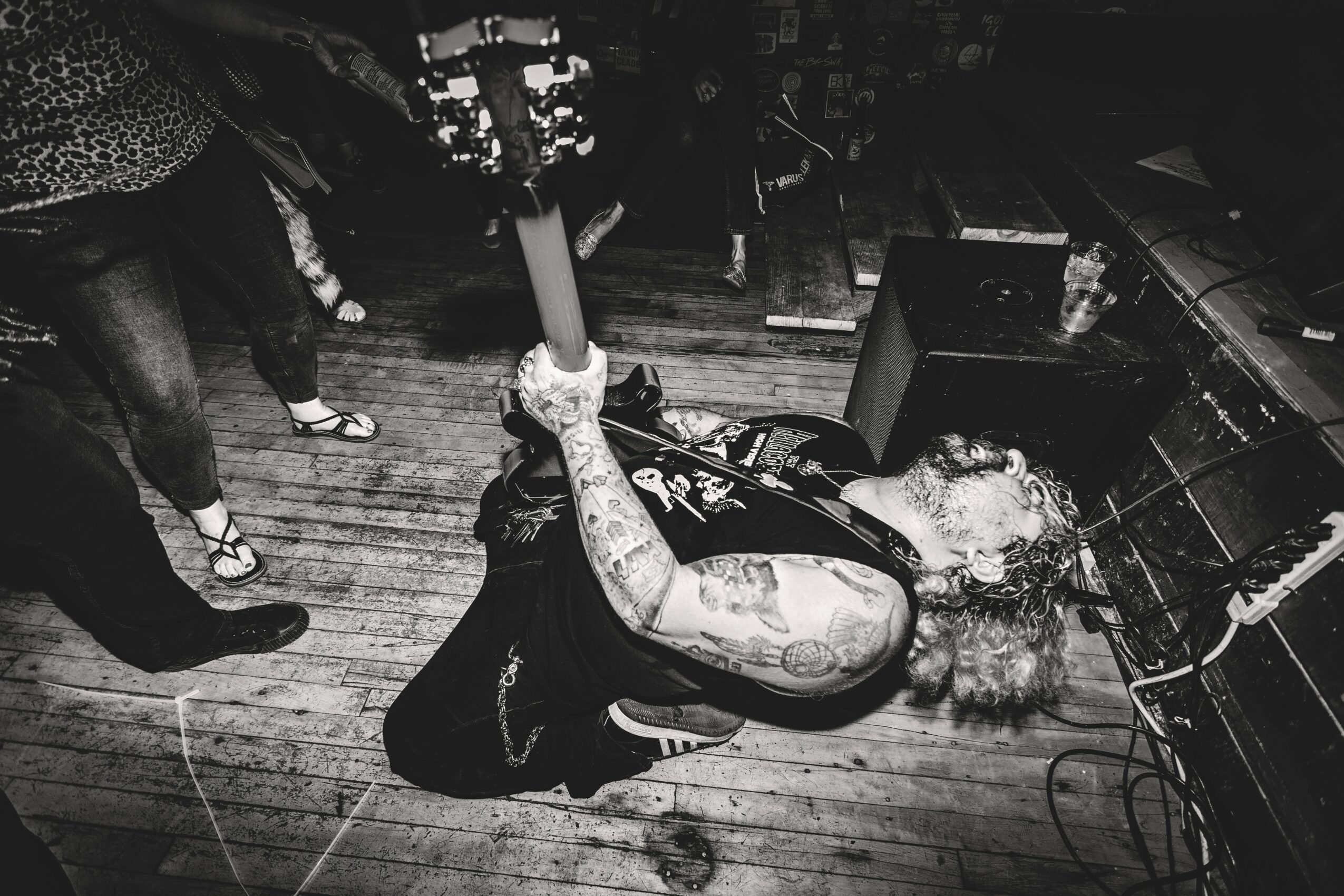 Person with curly hair and tattoos lying on the floor, holding a microphone, in a music venue or stage setting.