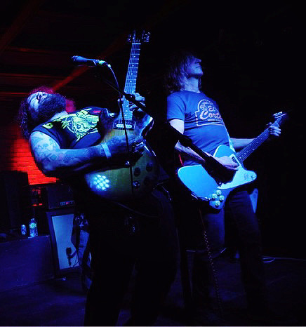 Two musicians perform on stage, one playing guitar and singing, the other playing an electric guitar, under blue and red lighting.