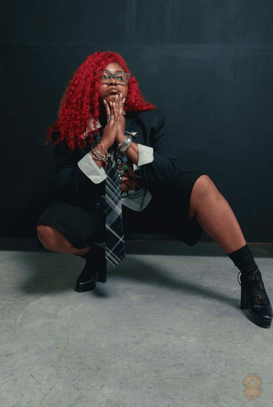 Person with curly red hair squatting against a dark background, wearing a blazer, tie, and boots, with hands clasped.