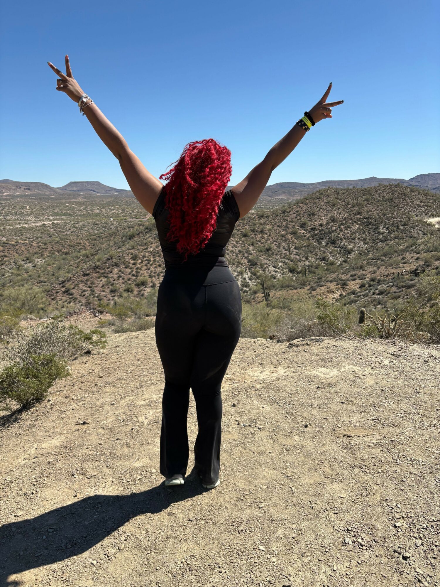 Person with red hair standing outdoors on a dirt path, arms raised with peace signs, landscape background, clear blue sky.