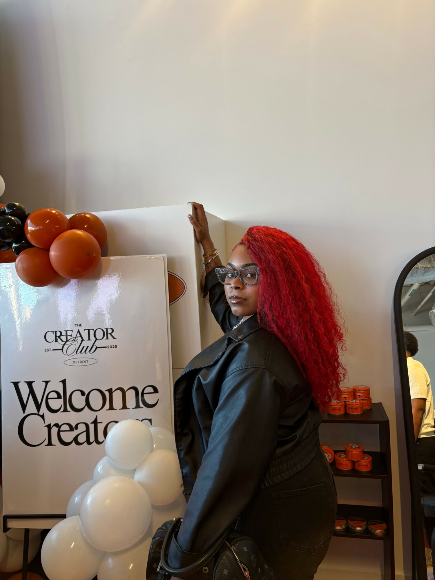 Woman with red hair in black jacket standing near a white wall with a welcome sign and decorative white and brown spheres.