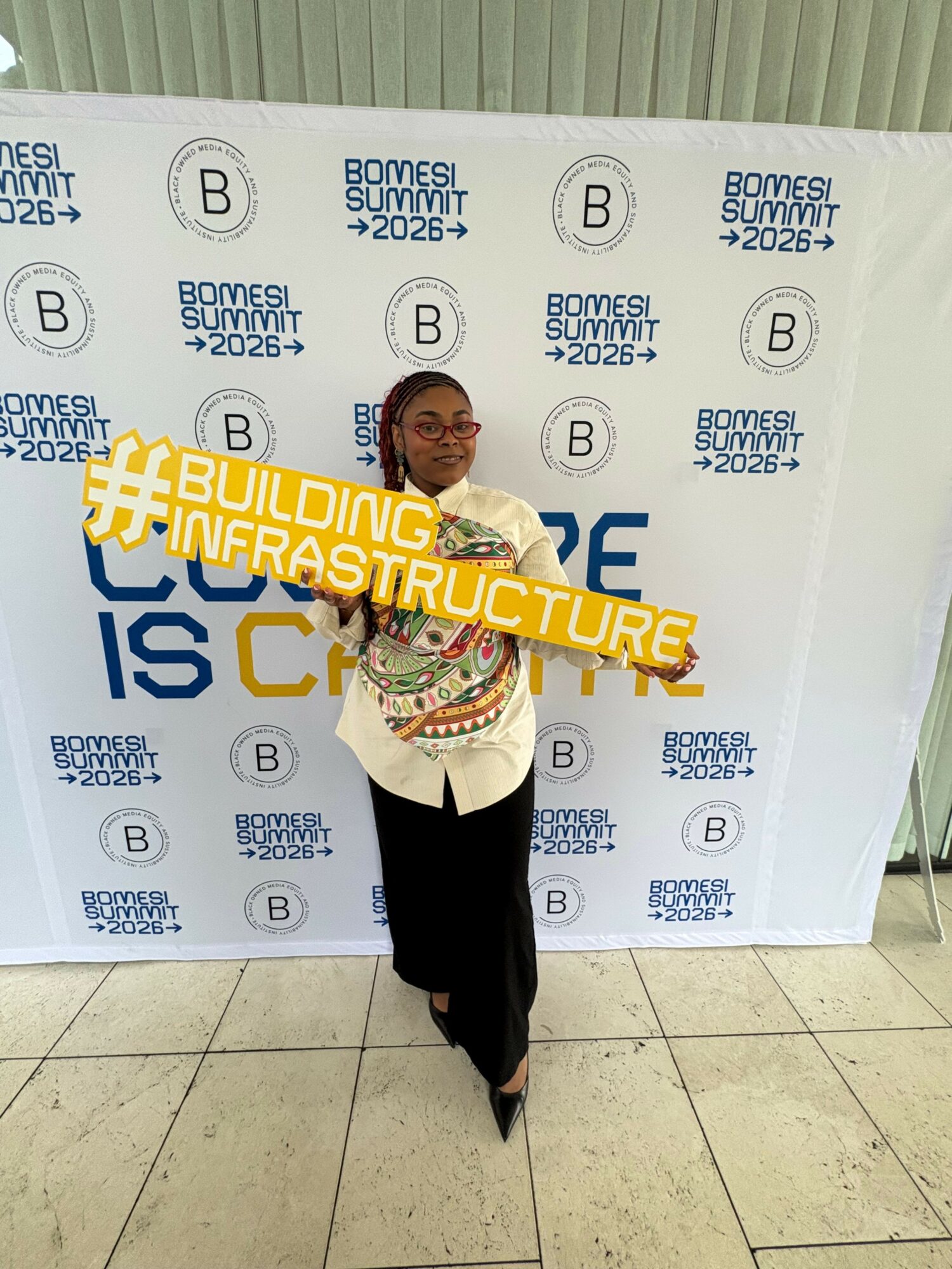 Person holding a yellow hashtag sign in front of a white backdrop with blue text and logos, standing on tiled floor.