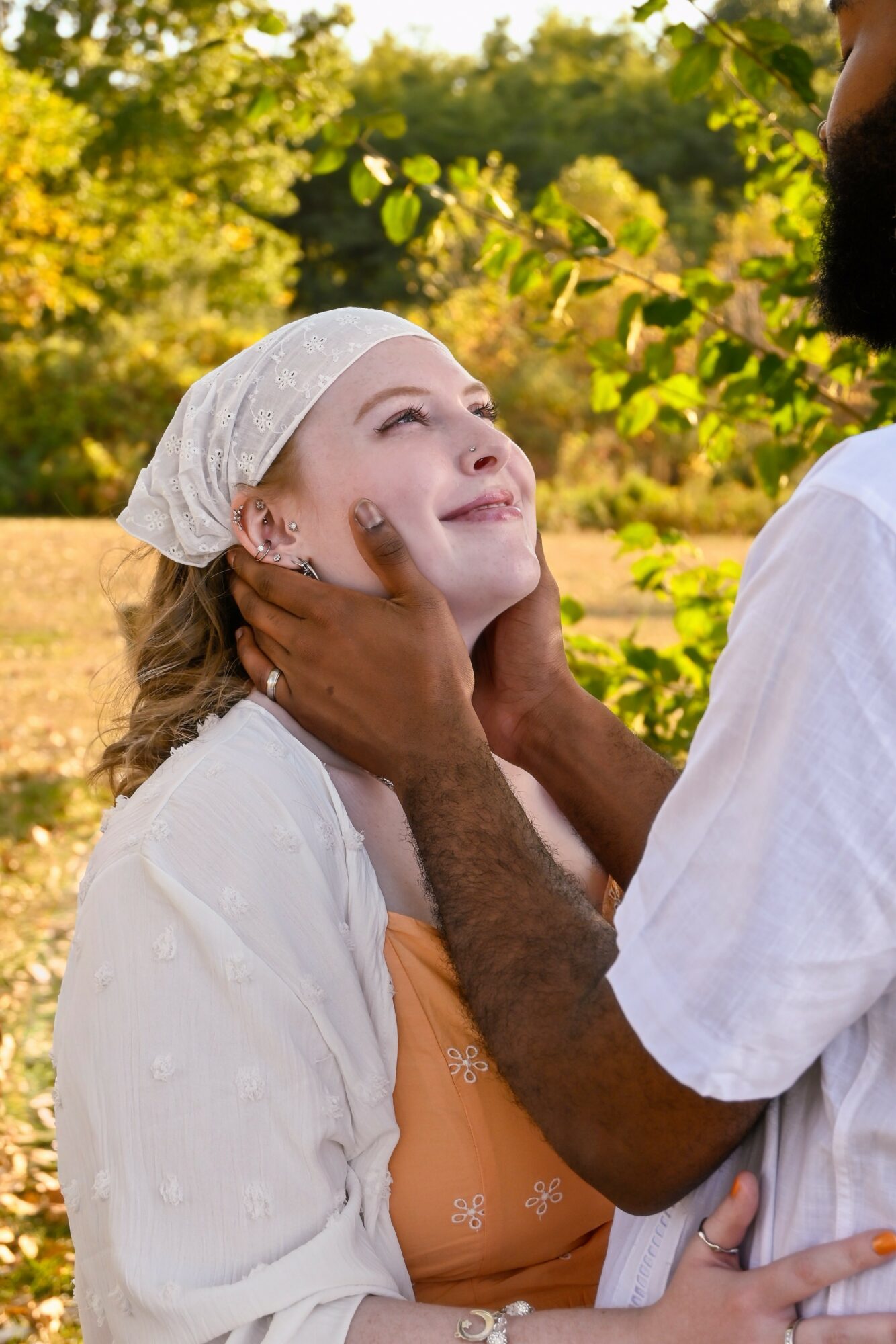Person gently holds a young girl's face outdoors, both smiling, surrounded by green trees and sunlight.