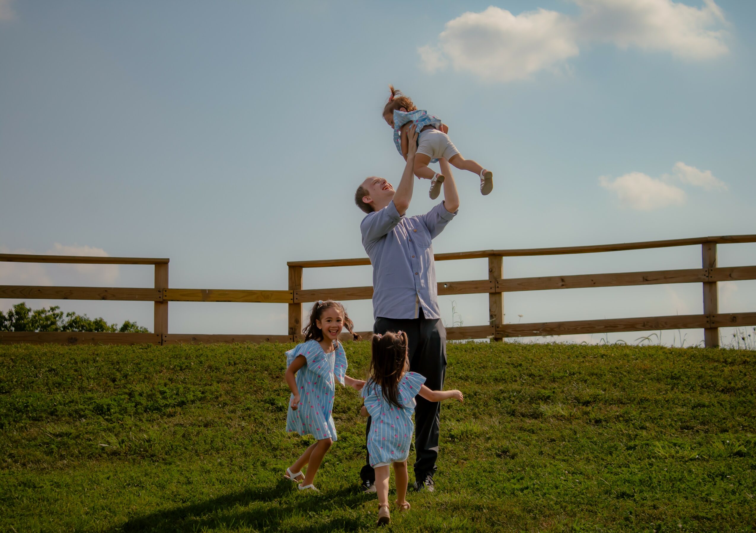 Man lifts girl in the air while two girls watch on grassy hill with fence, sky, and clouds in background.