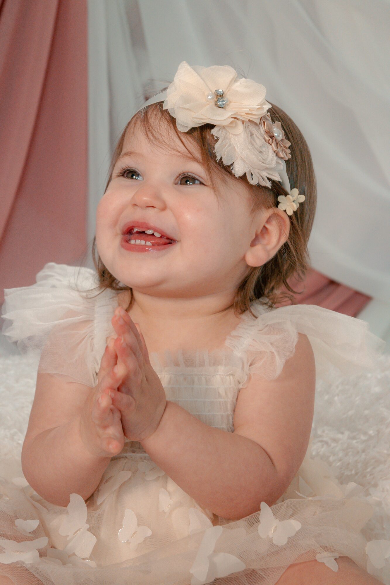 Smiling young girl with a floral headband, hands clasped, wearing a light-colored dress with tulle, in front of pastel-colored drapes.