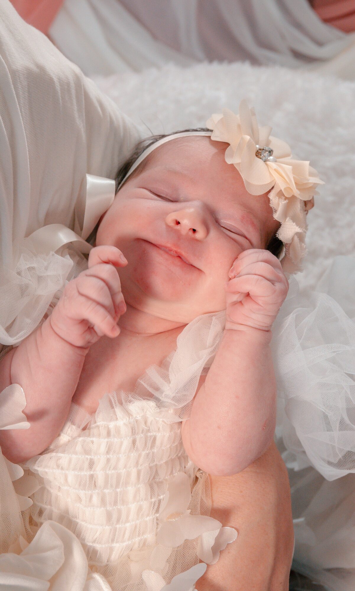 Smiling baby girl with a floral headband, lying on a soft surface, wearing a light-colored dress, hands near face.