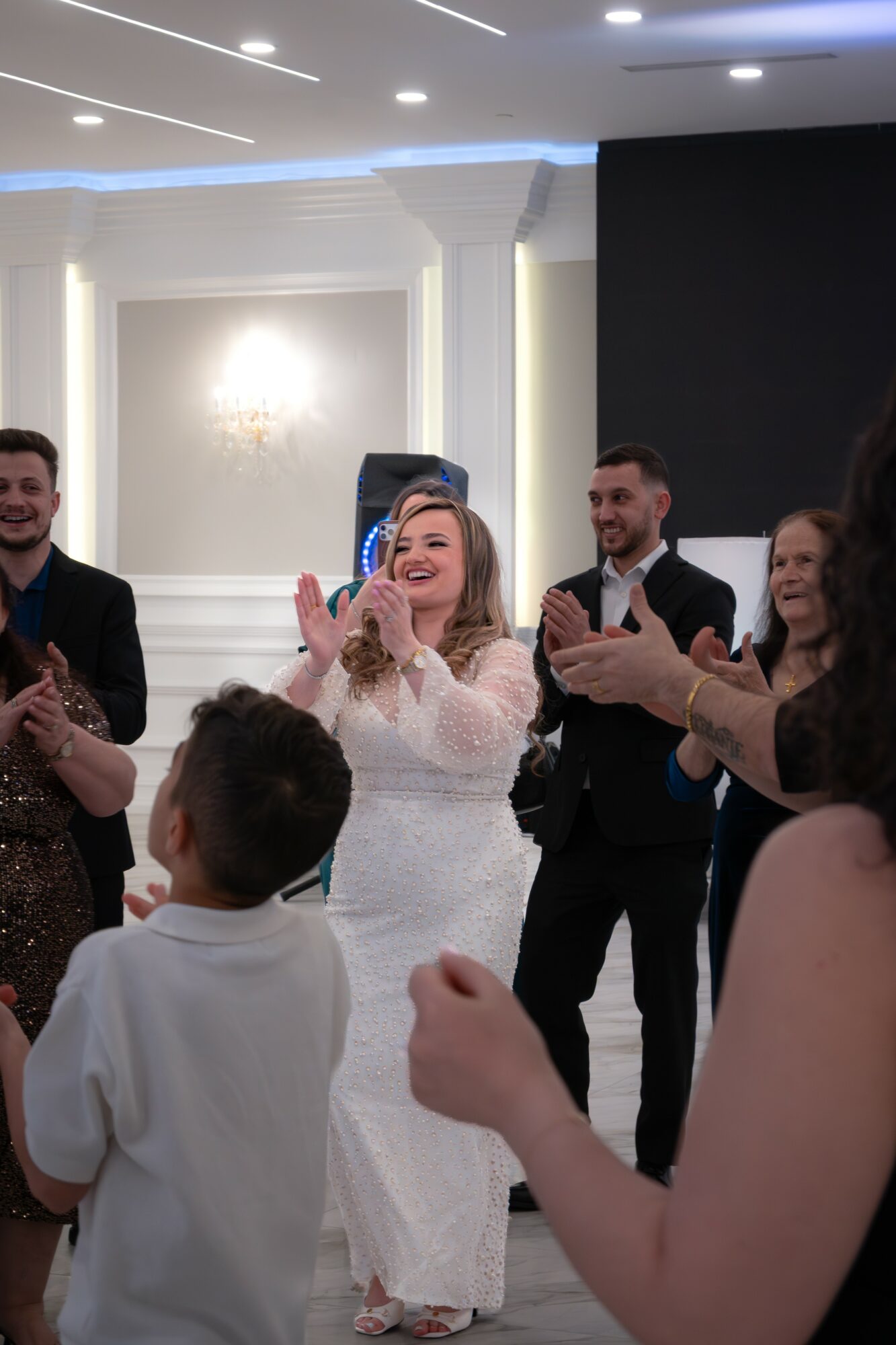 Group of people celebrating indoors, woman in white dress clapping, others smiling and clapping, elegant room with white walls and lighting.