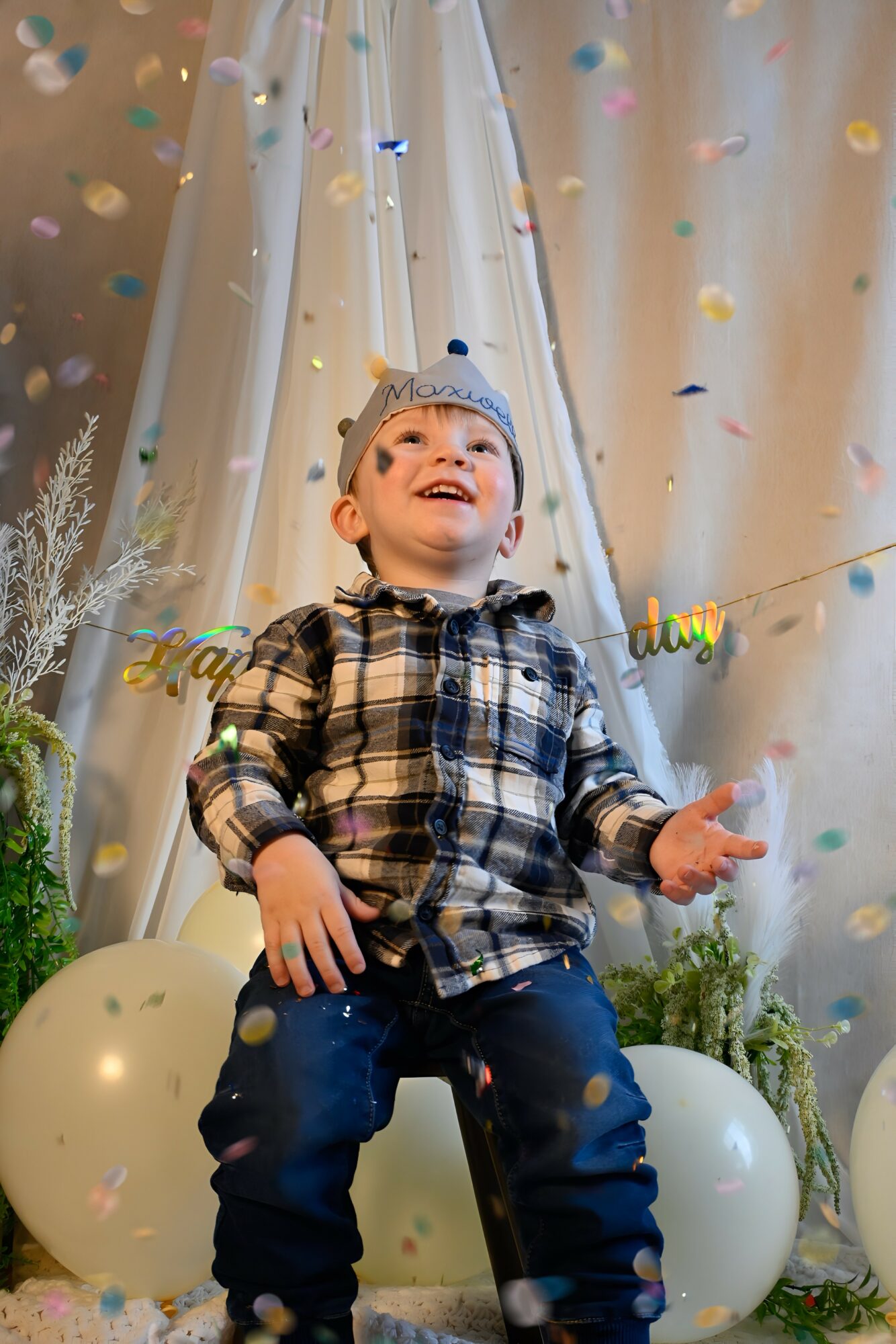 Child in plaid shirt and birthday hat smiling with confetti falling around him, standing between balloons and plants.