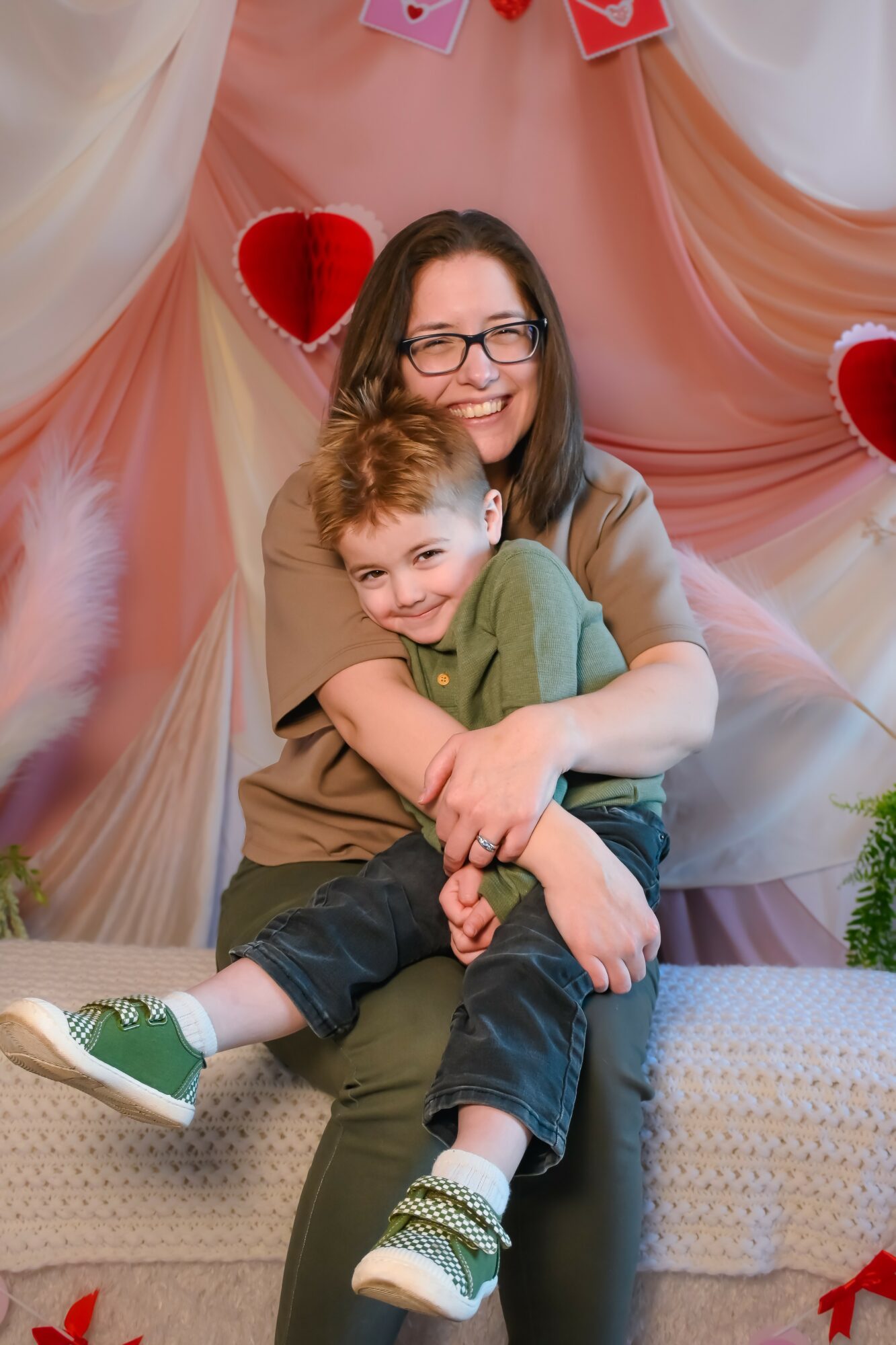 Woman with glasses hugging a smiling child in front of a decorated backdrop with hearts and drapes.