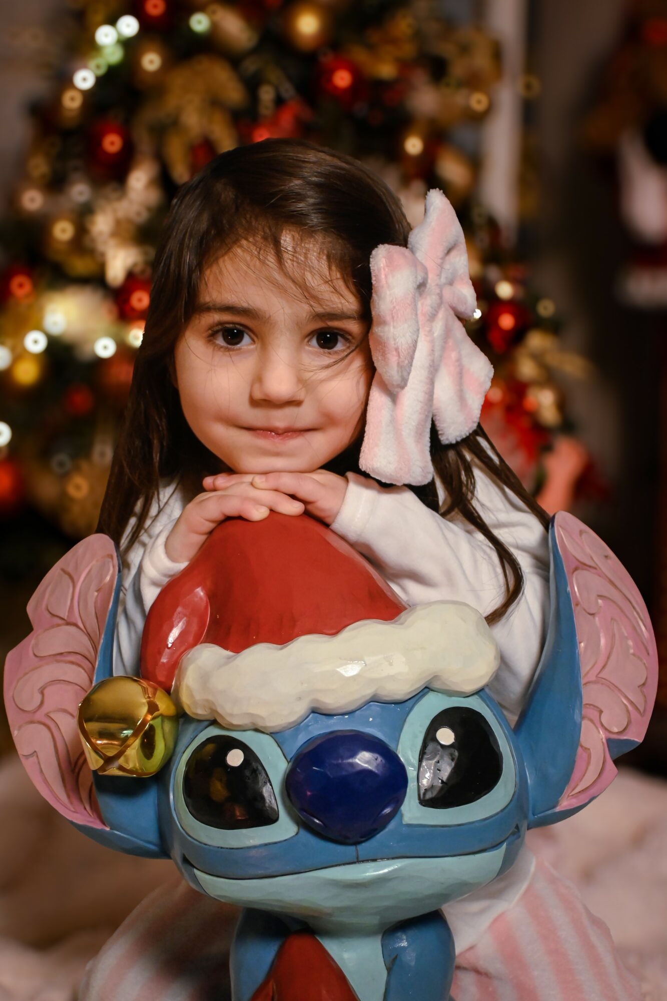 Young girl with a pink bow in her hair, resting her chin on a colorful toy, in front of a decorated Christmas tree.