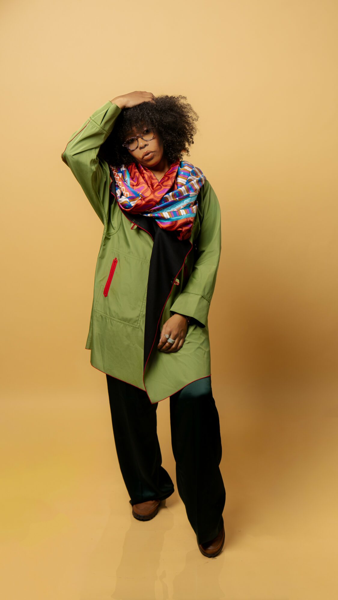 Person with curly hair wearing a green coat, colorful scarf, black pants, standing against a beige background.