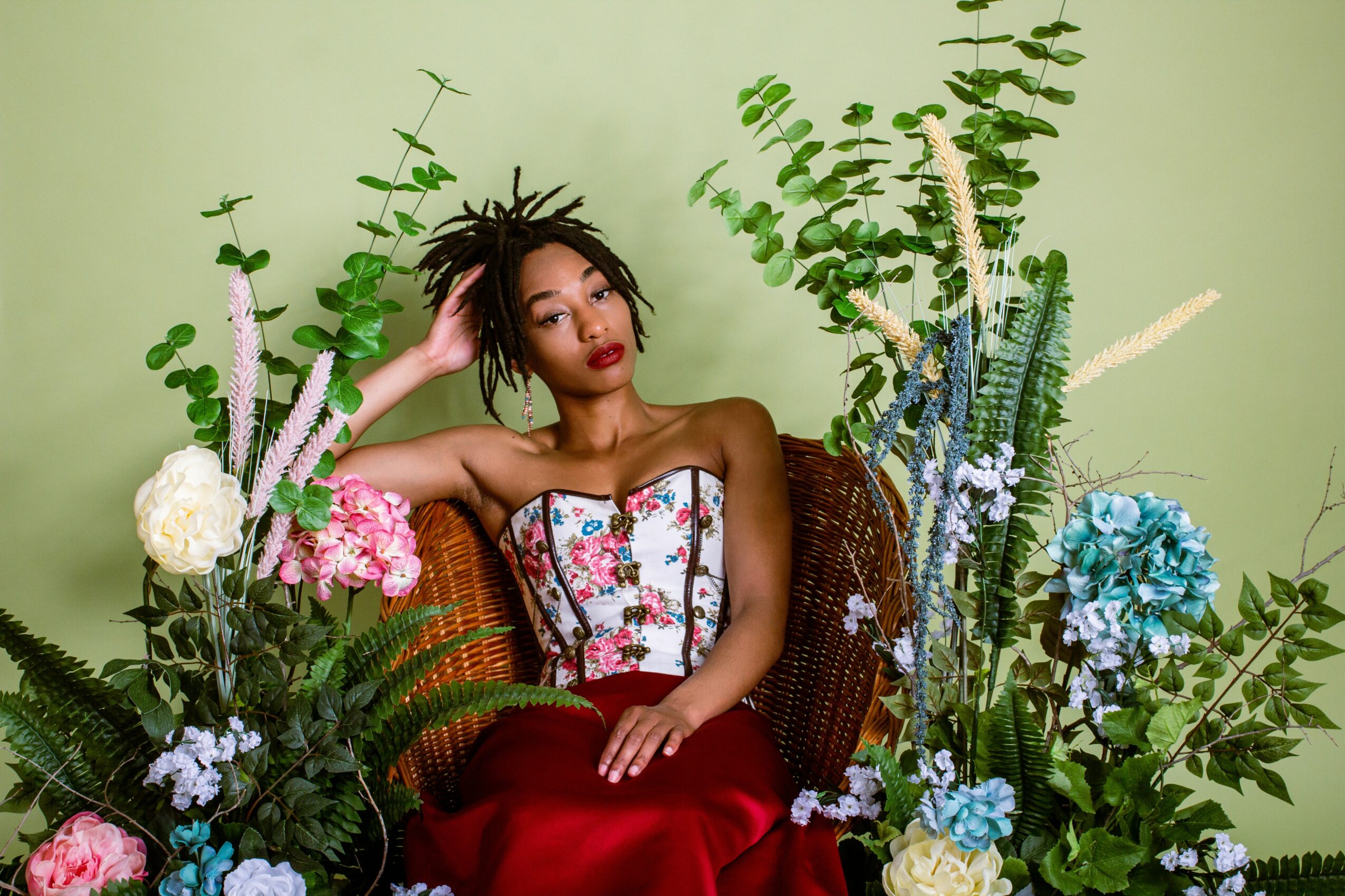 Woman with short hair and red lipstick sitting among colorful flowers and greenery, wearing a strapless dress with floral pattern.