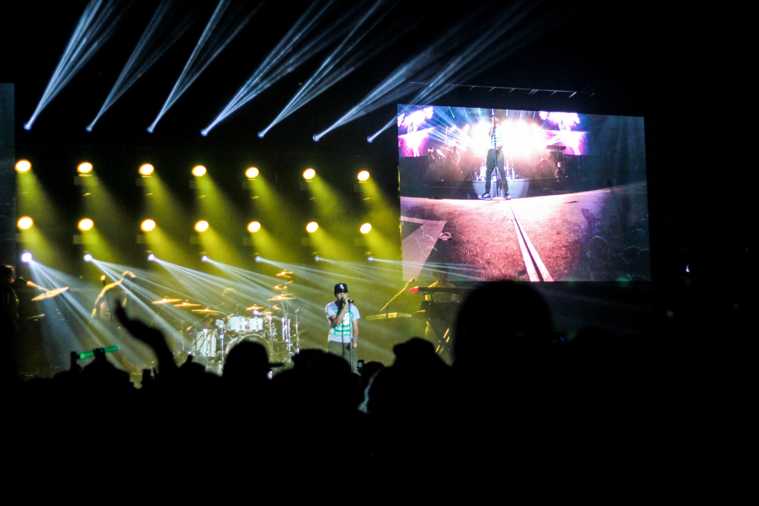 Stage with bright yellow and white lights, large screen displaying a person, audience silhouettes visible.