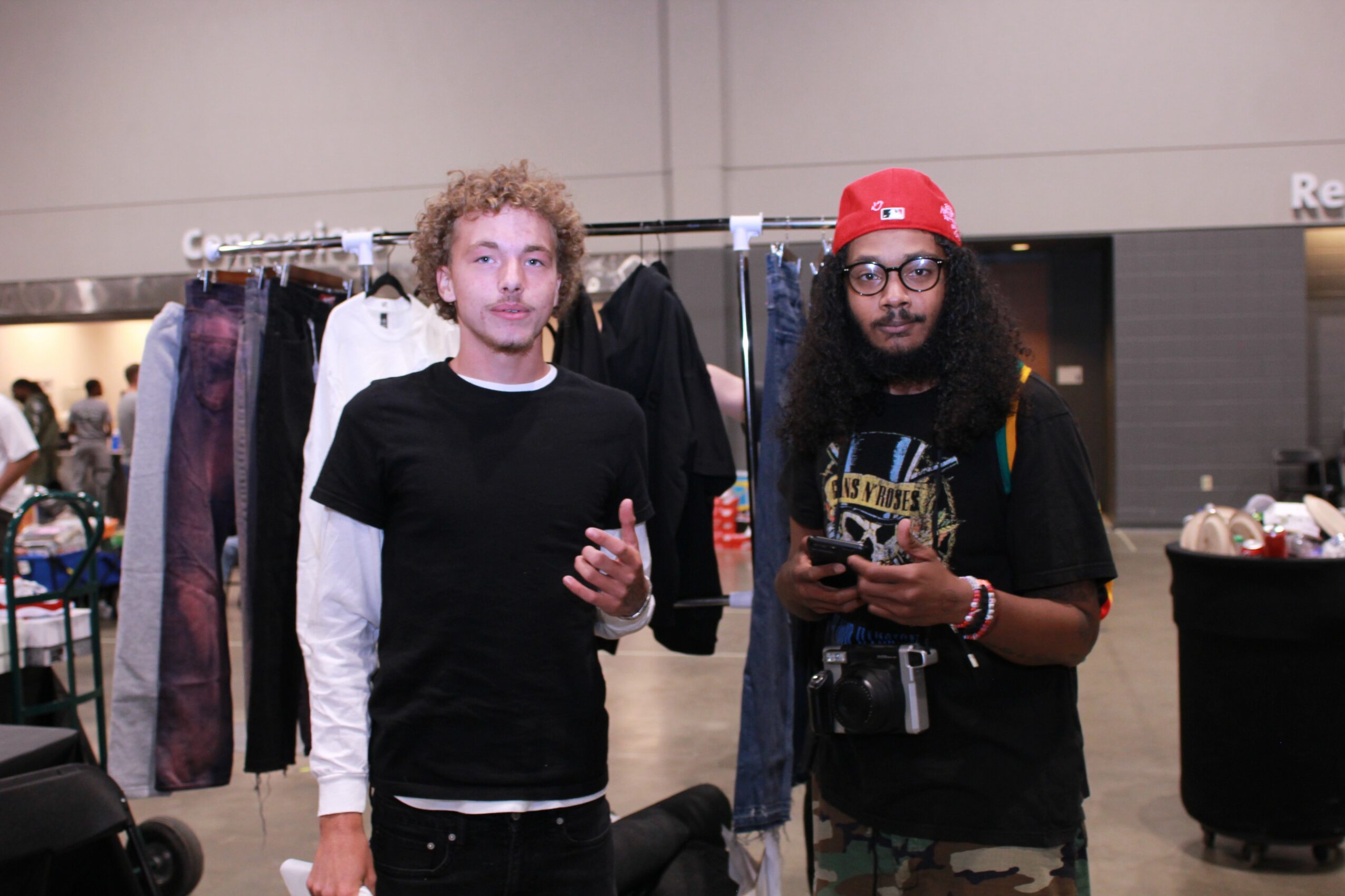 Two people standing in front of a clothing rack at an indoor event, one with curly hair and the other with long hair and a red cap.