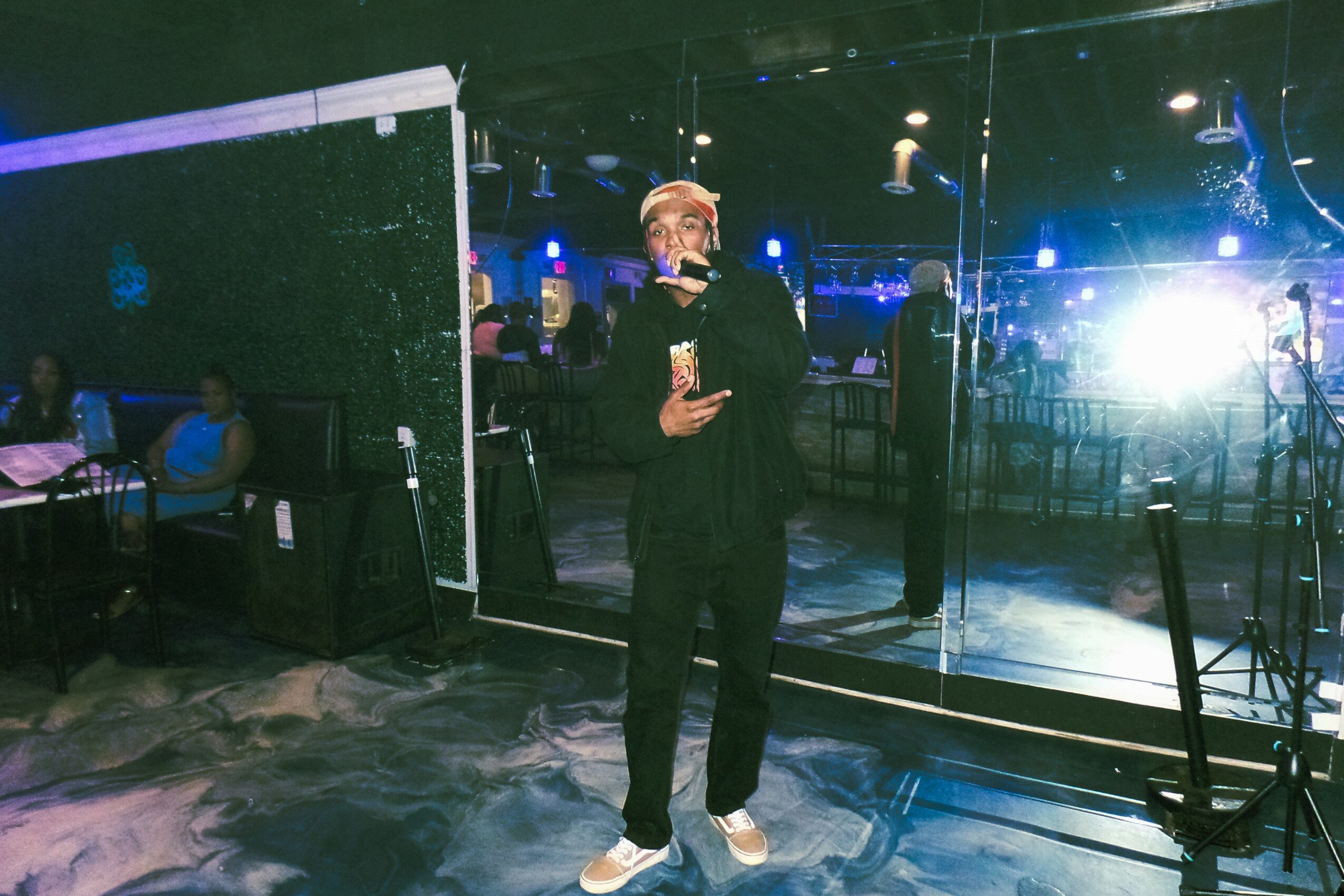 Person taking a mirror selfie in a dimly lit venue with blue and purple lighting, people in the background, and a reflective wall.