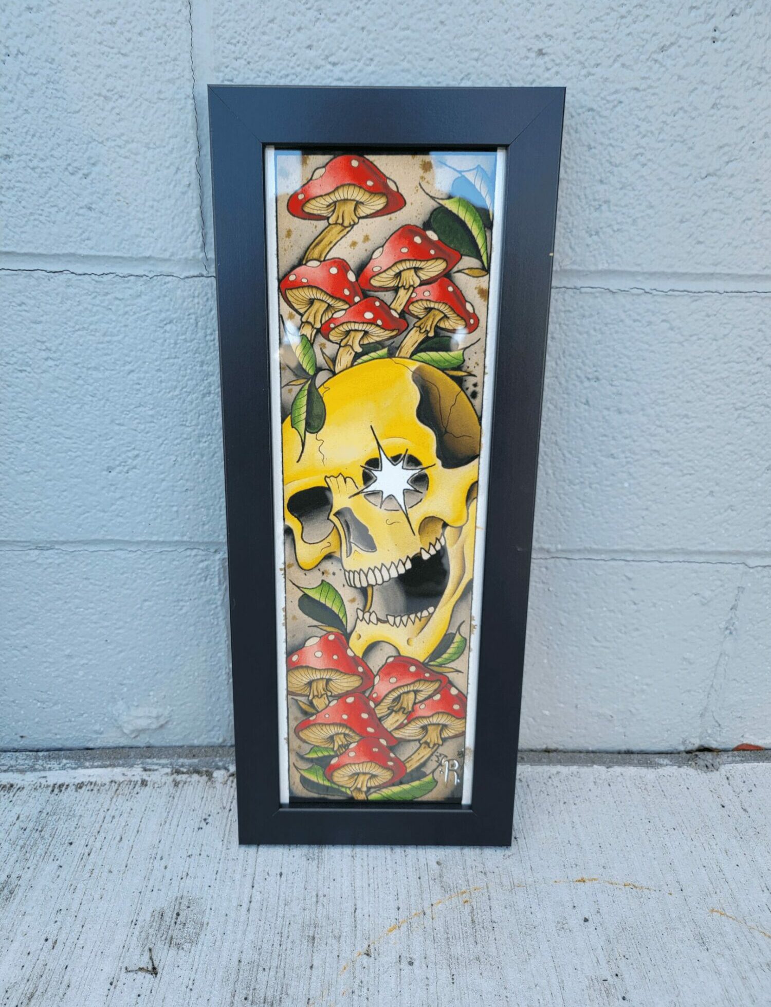Framed artwork featuring a yellow skull with a star-shaped eye, surrounded by red mushrooms with white spots, on a colorful background.