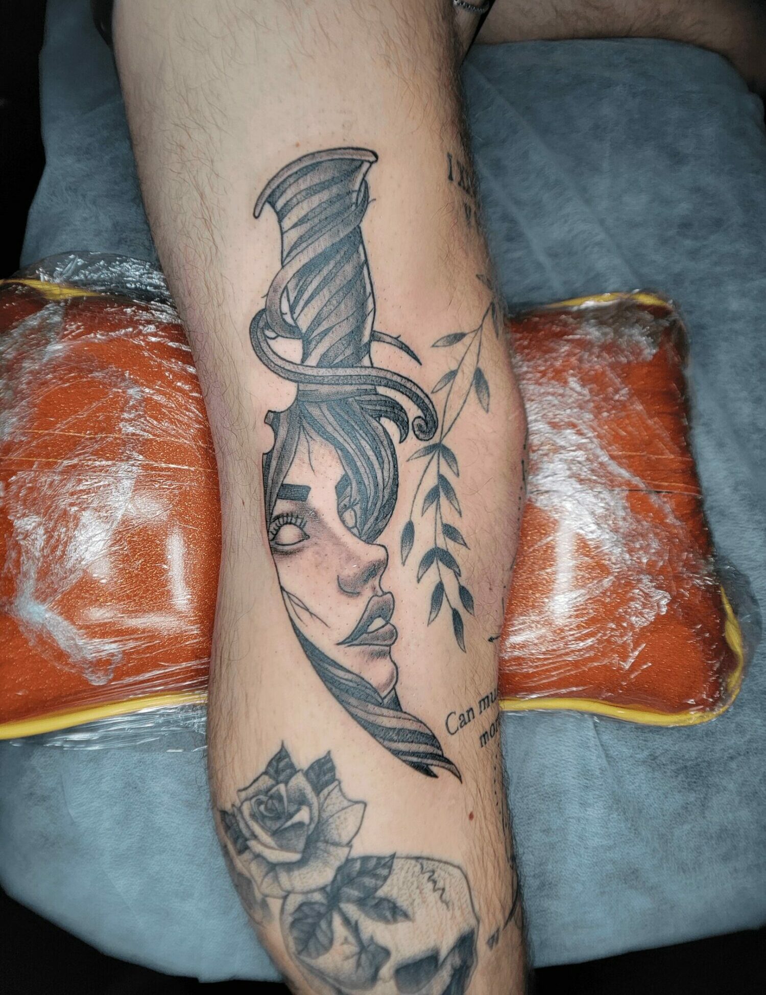 Tattoo of a woman's face with flowing hair and leaves on a person's arm, with a rose below. The arm rests on an orange cushion.