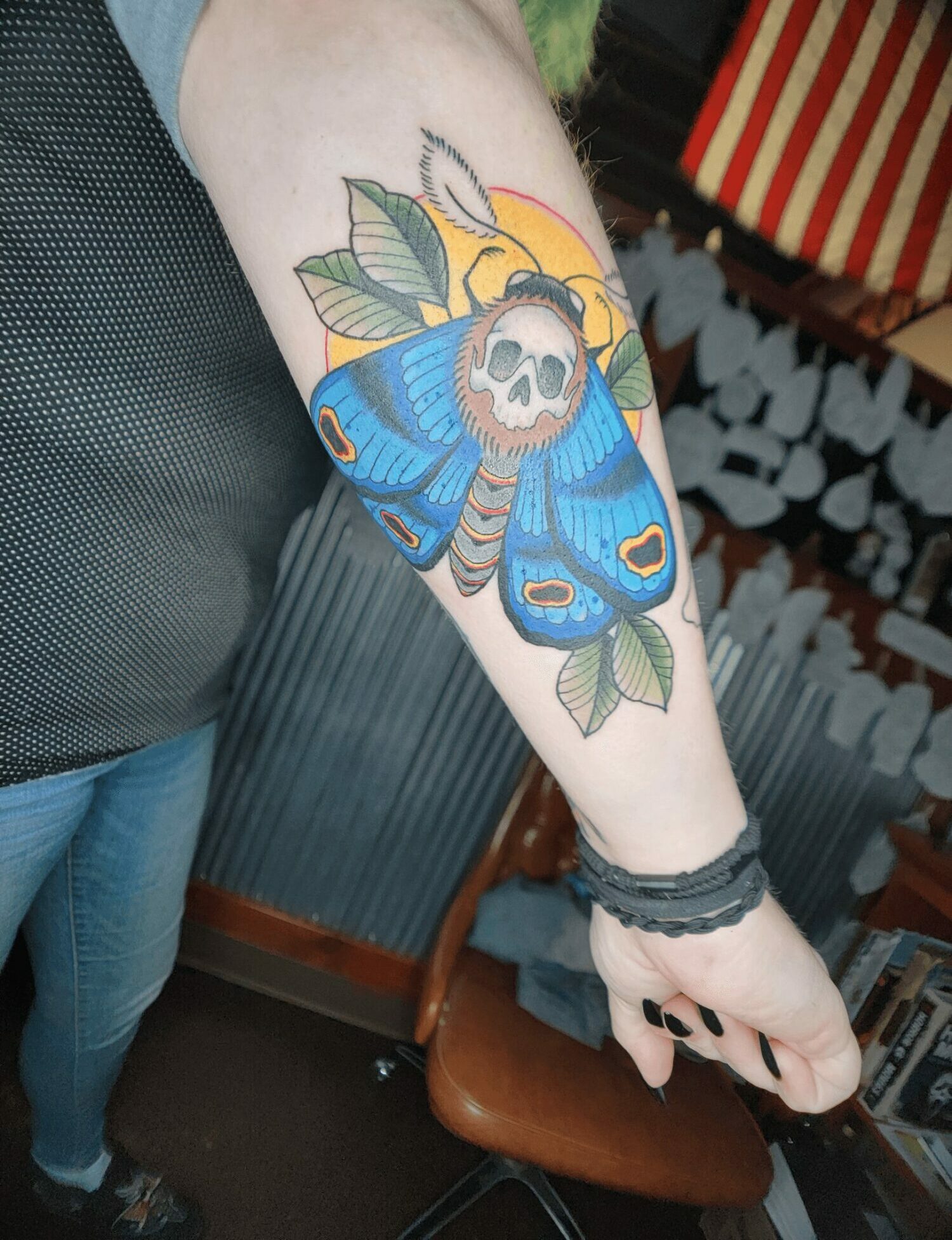 Tattoo of a blue butterfly with a skull at the center, surrounded by leaves and a sun, on person's arm.