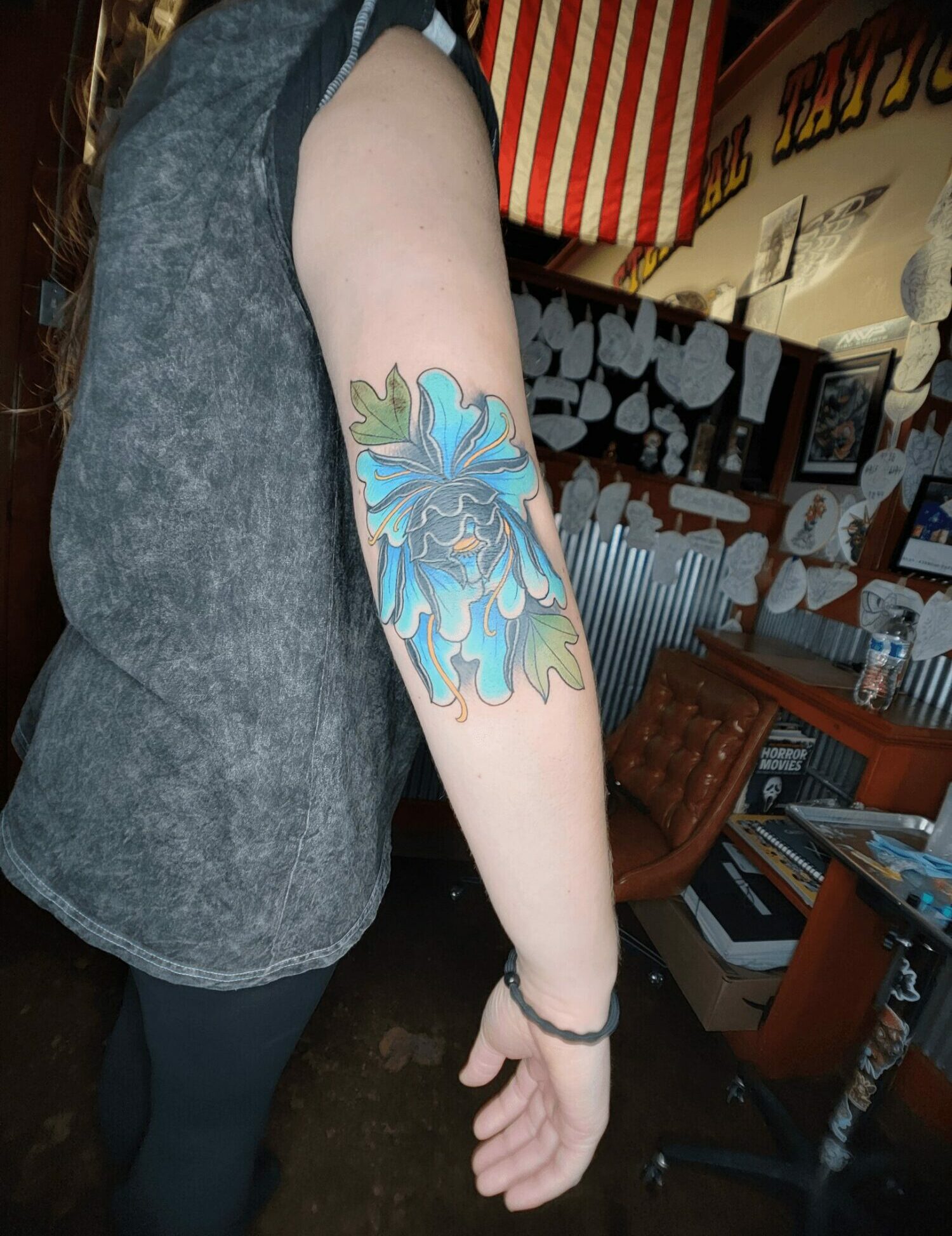 Person's arm with a colorful tattoo of a blue and purple flower with green leaves, indoors setting.