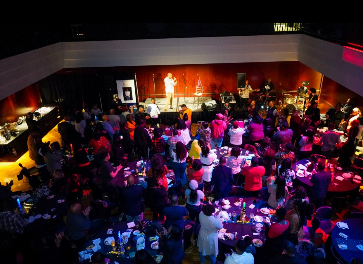 Audience watching a live performance on stage in a large, dimly lit venue with colorful lighting and tables.