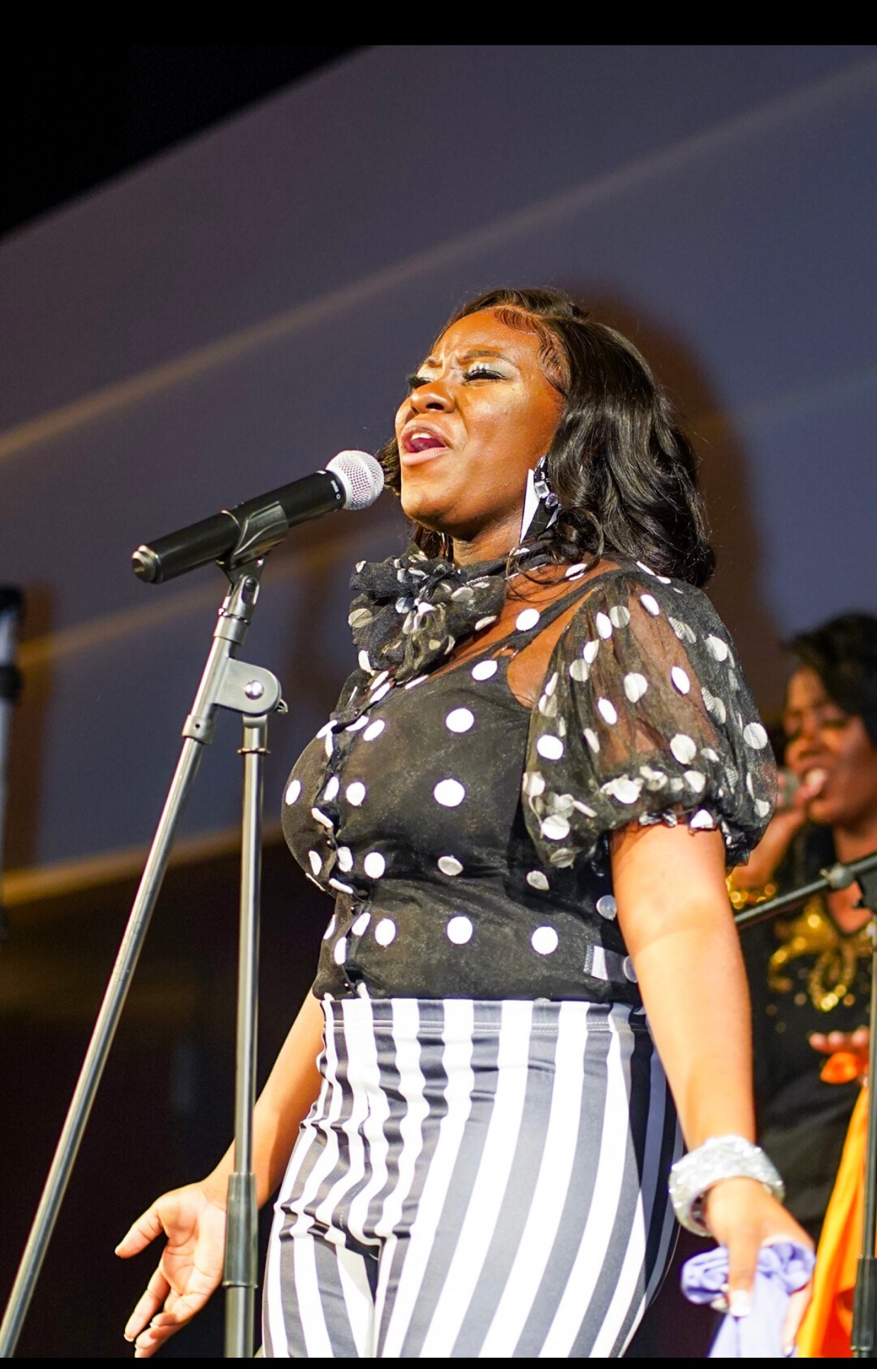 Woman singing into microphone on stage, wearing polka dot top and striped pants, with a smiling person in background.