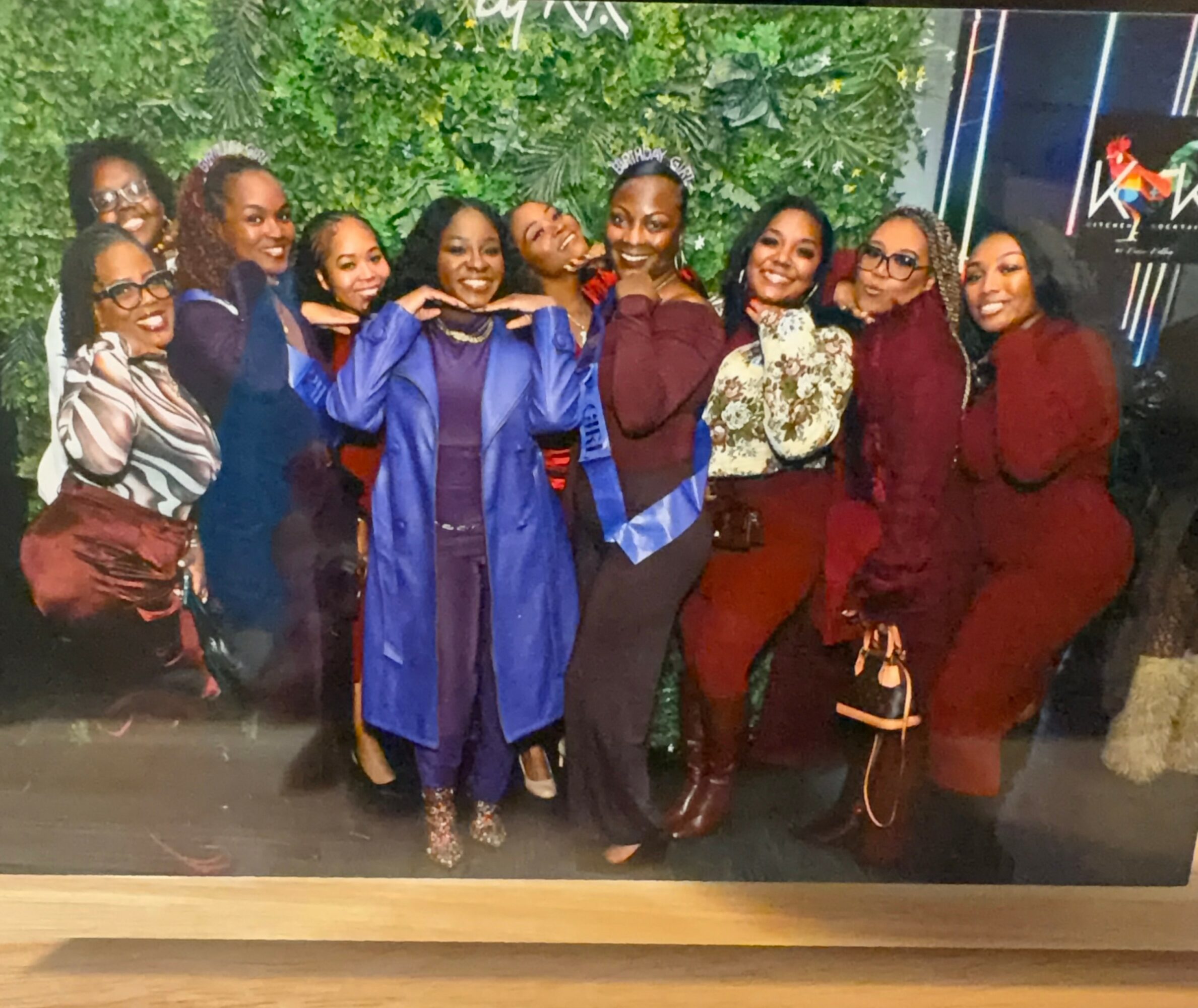 Group of women celebrating graduation, wearing formal and colorful outfits, smiling, with greenery and a building in the background.