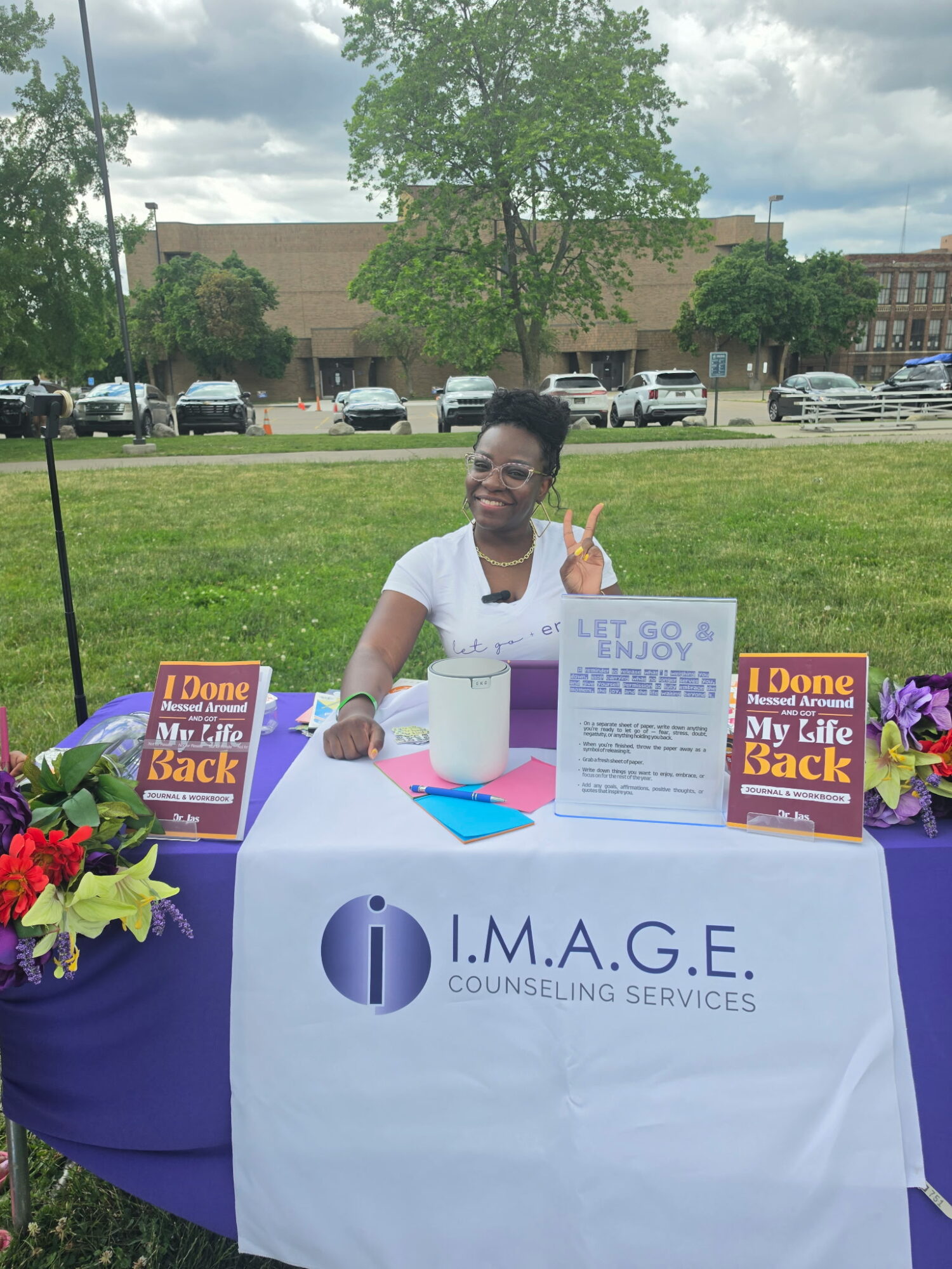 Smiling woman standing behind a table with counseling service signs outdoors, with trees and parked cars in background.
