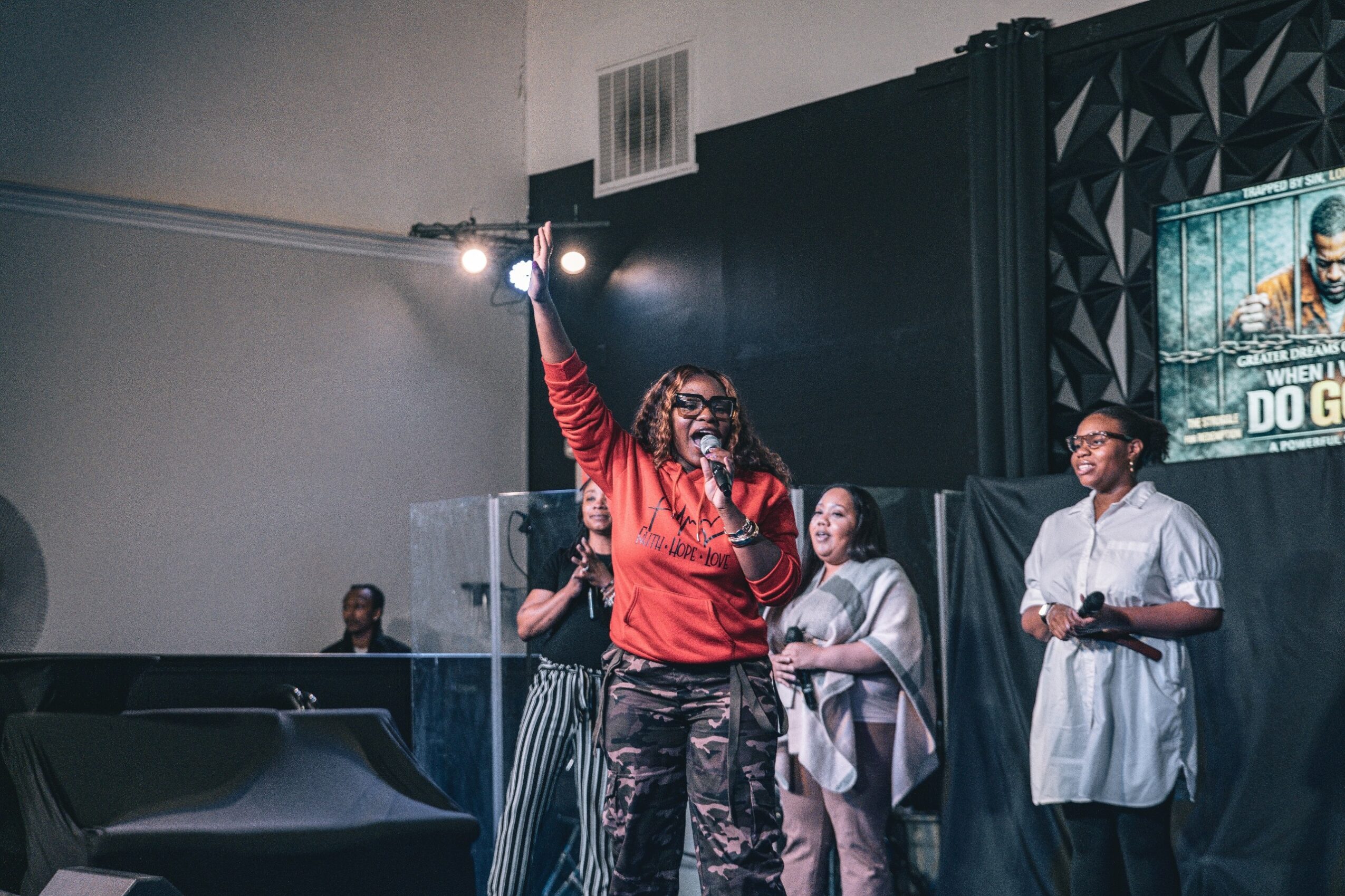 Woman with glasses and red jacket speaking into microphone on stage, with three people behind her, in a dark room.