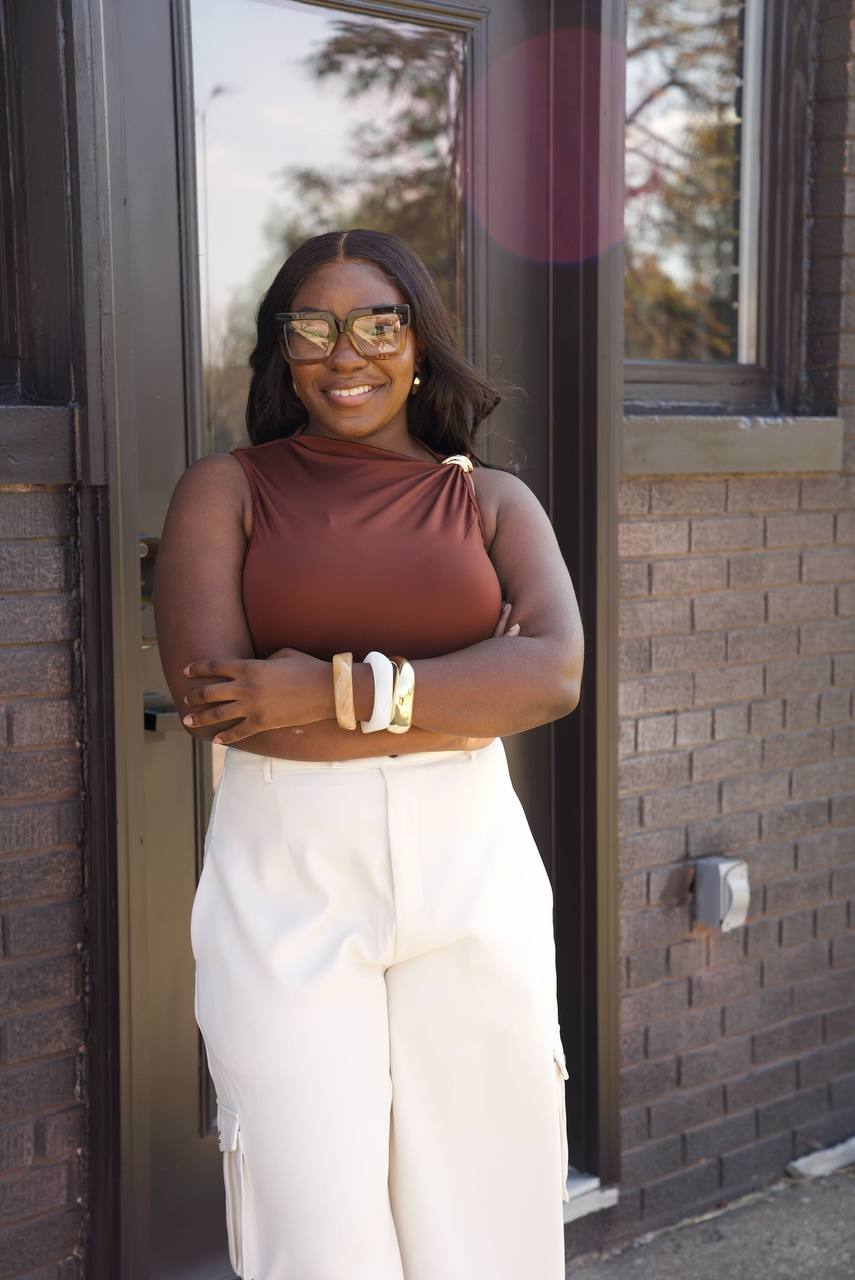 Smiling woman with glasses and dark hair stands outside a building with brick wall and windows, arms crossed, wearing a brown top and white pants.