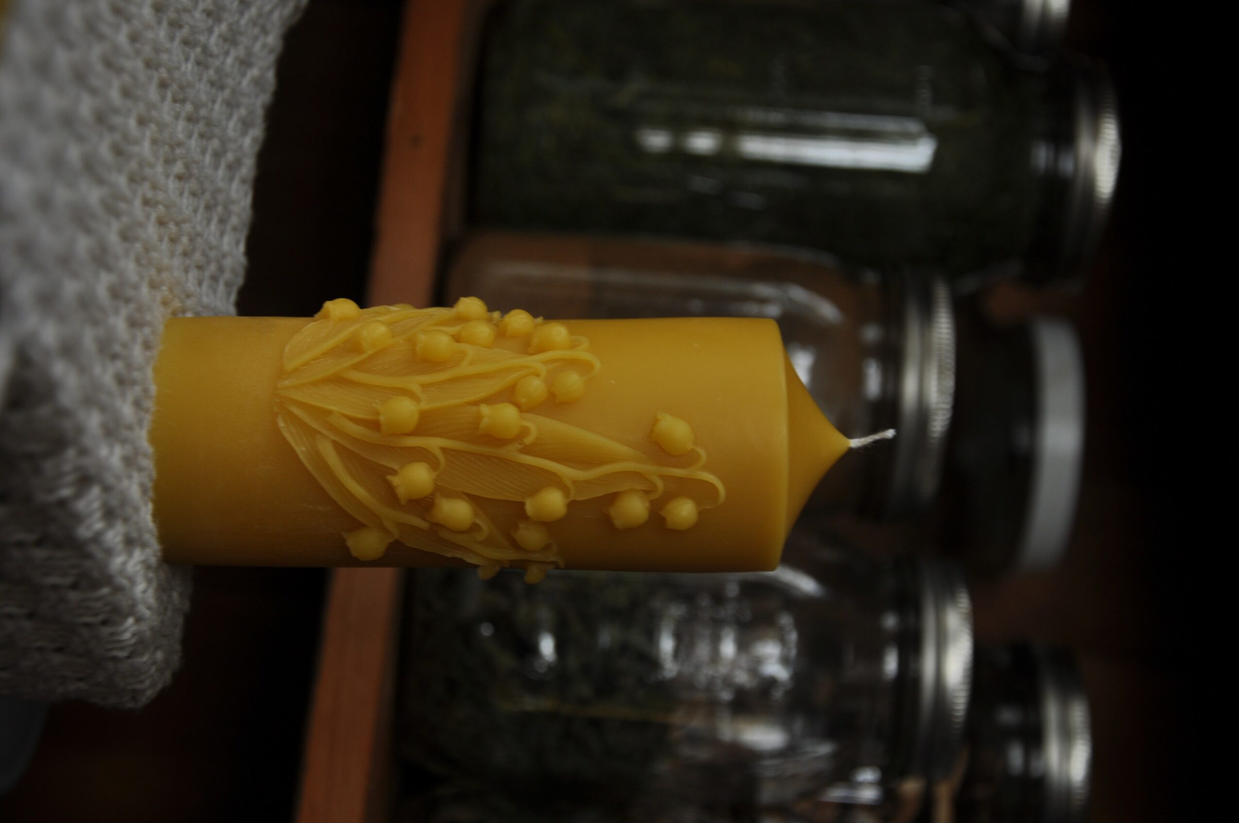 Yellow candle with decorative wax drips, placed on a textured gray surface, with glass jars in the background.