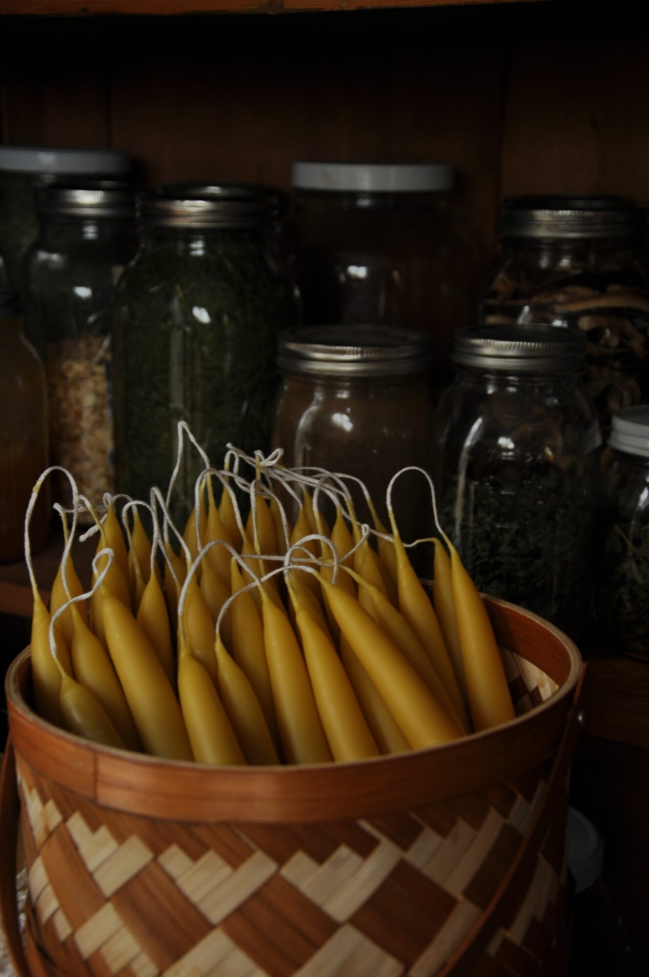 A woven basket filled with yellow candles with wicks, placed on a shelf with jars behind it.