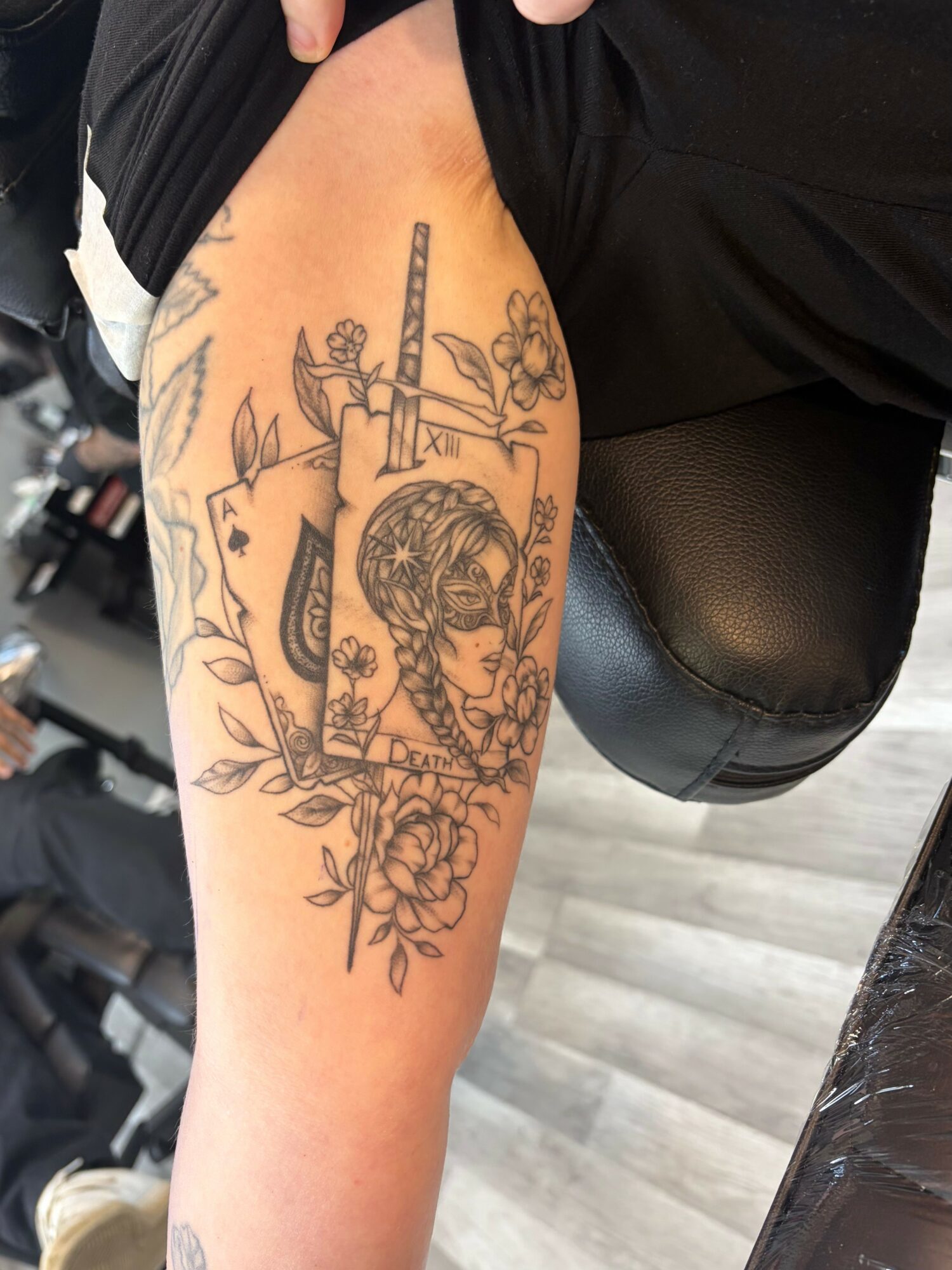 Tattoo of a woman’s face with floral and sword elements on thigh, black ink, detailed line work.