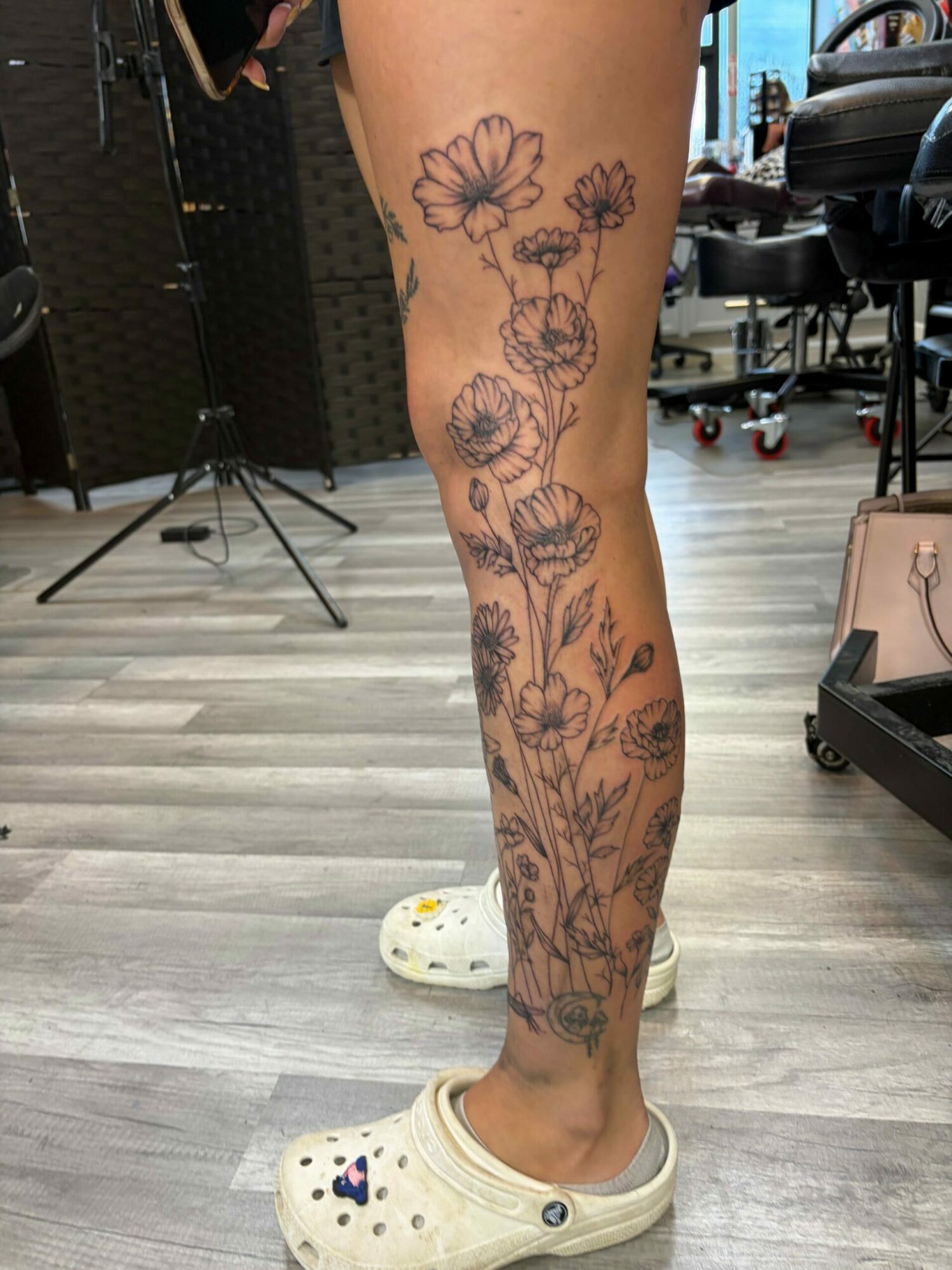 Person's leg with floral tattoo, wearing Crocs, in a room with wooden flooring and furniture.