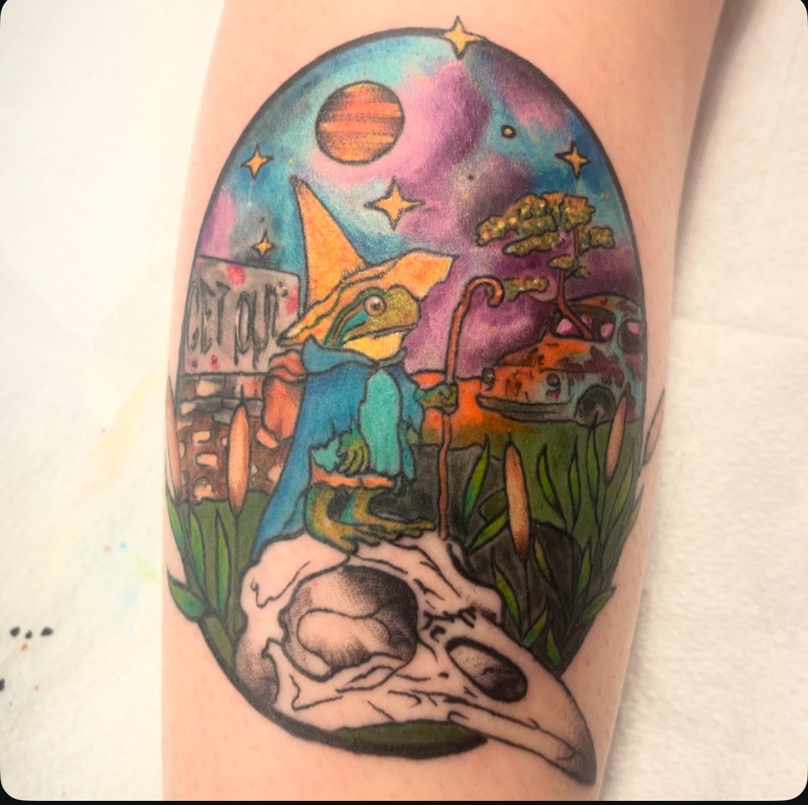 Tattoo of a frog wearing a blue hoodie sitting on a skull, with a colorful cosmic background including planets and stars.