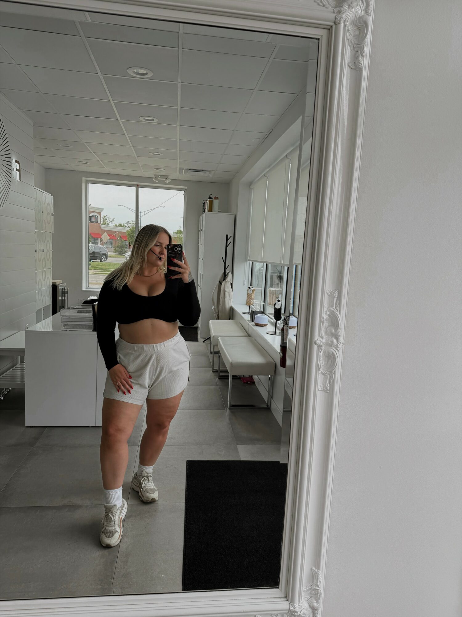 Woman taking a mirror selfie in a bright room with white furniture and a window, wearing a black top and white shorts.
