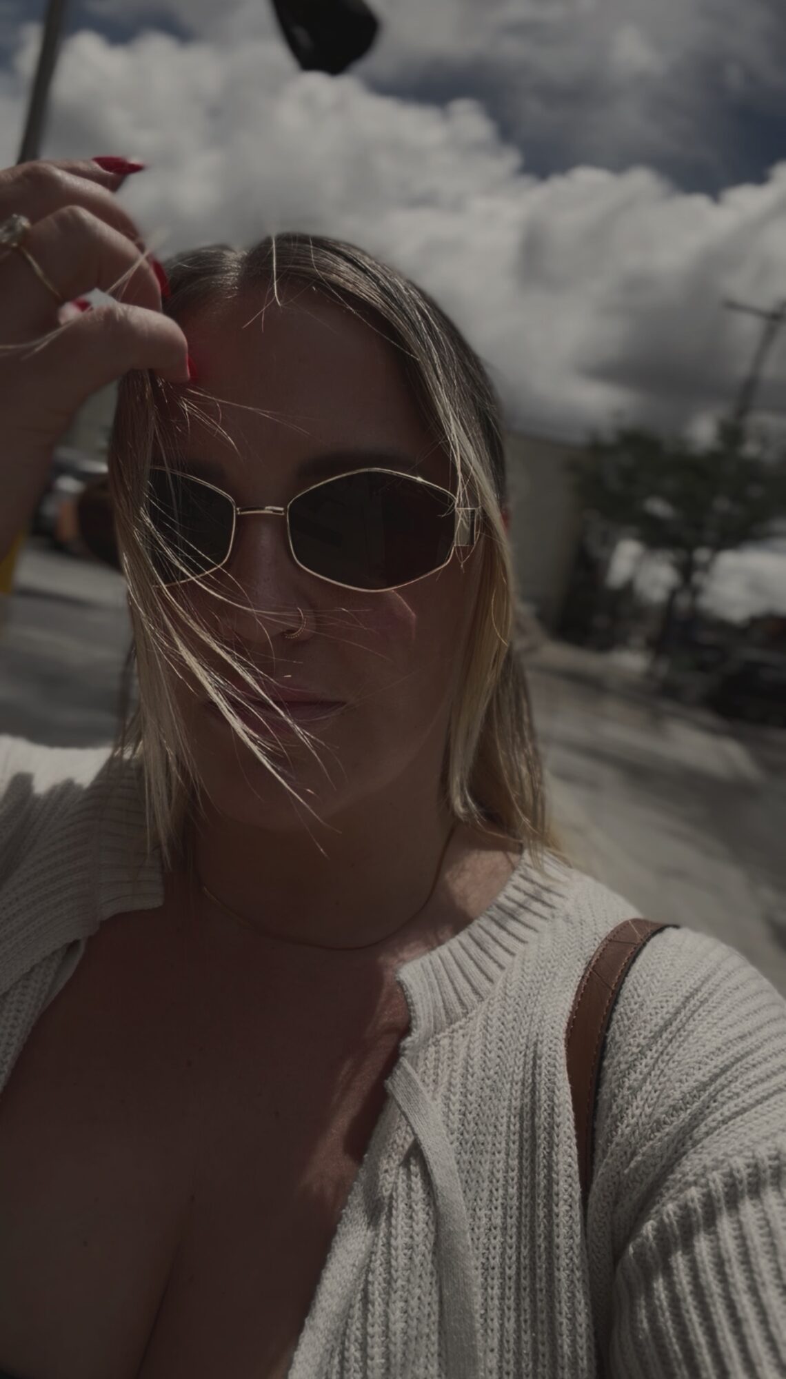 Woman wearing sunglasses and a light-colored top outdoors with cloudy sky background.